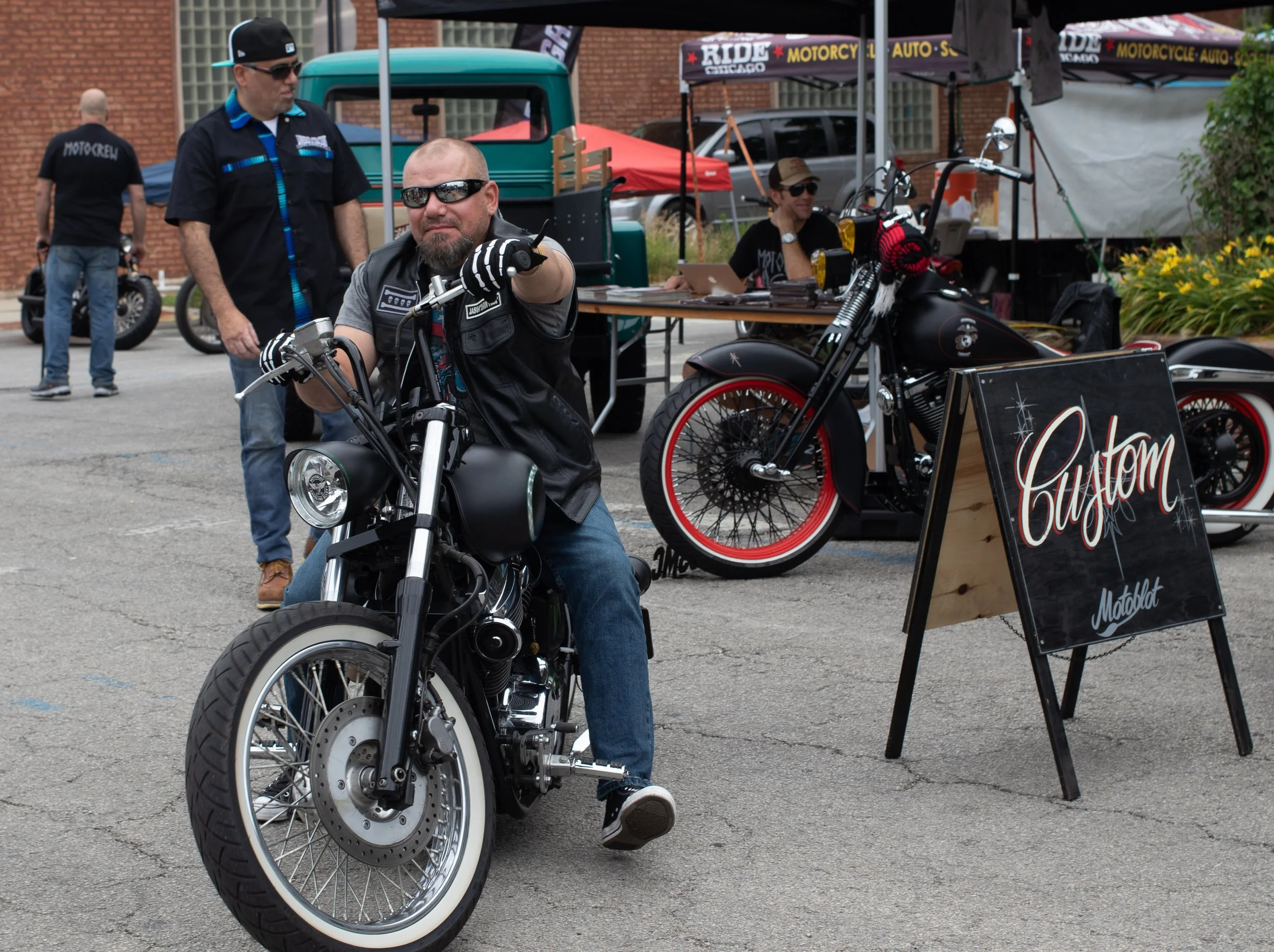 A man wearing sunglasses and a vest sitting on a black motorcycle at an outdoor event, with booths and people in the background.