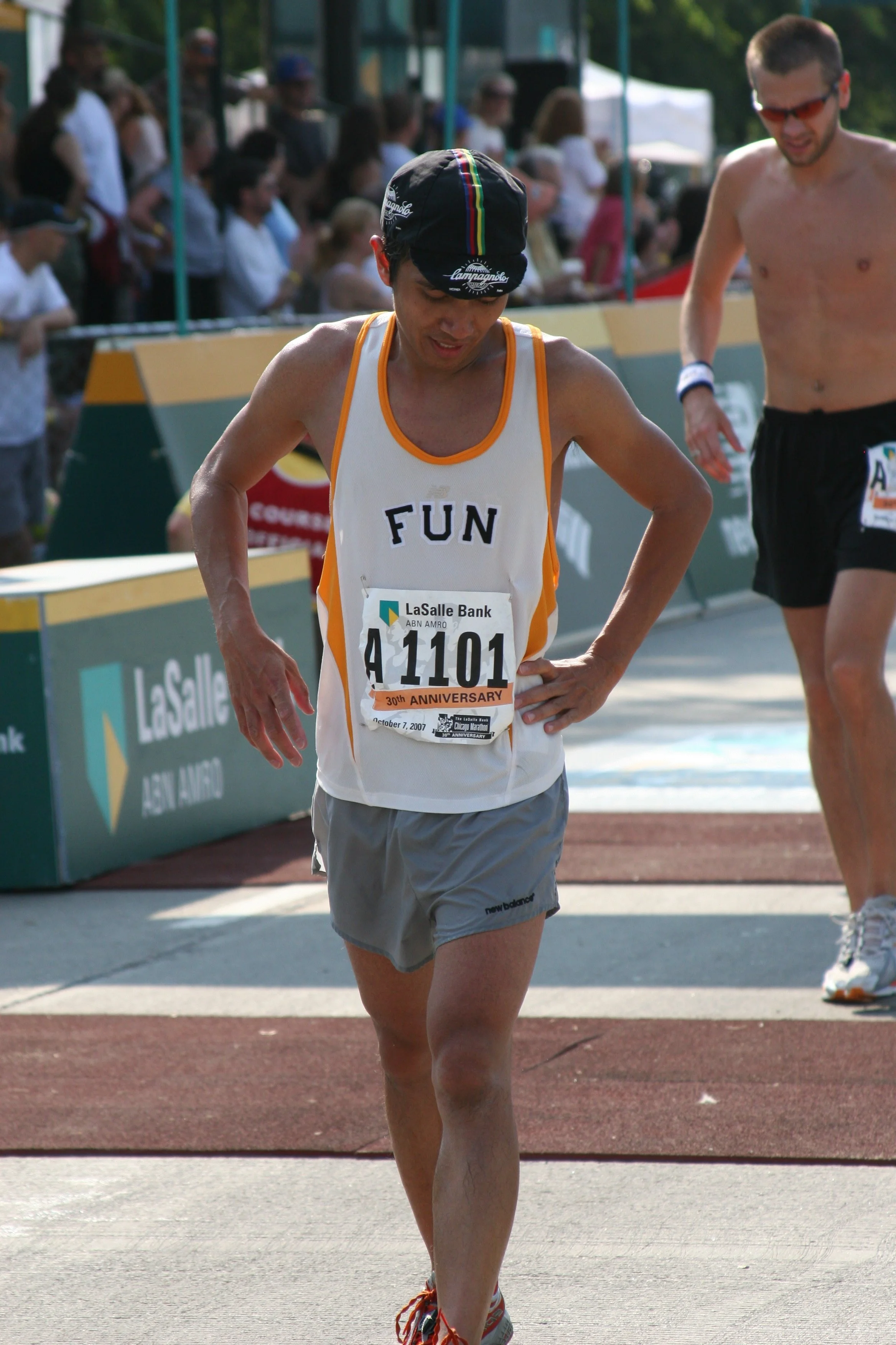 A man with a bib number 4101 participating in a marathon or running event, wearing a white tank top with orange trim, gray shorts, and a black cap. There is a crowd in the background and another runner to the right.
