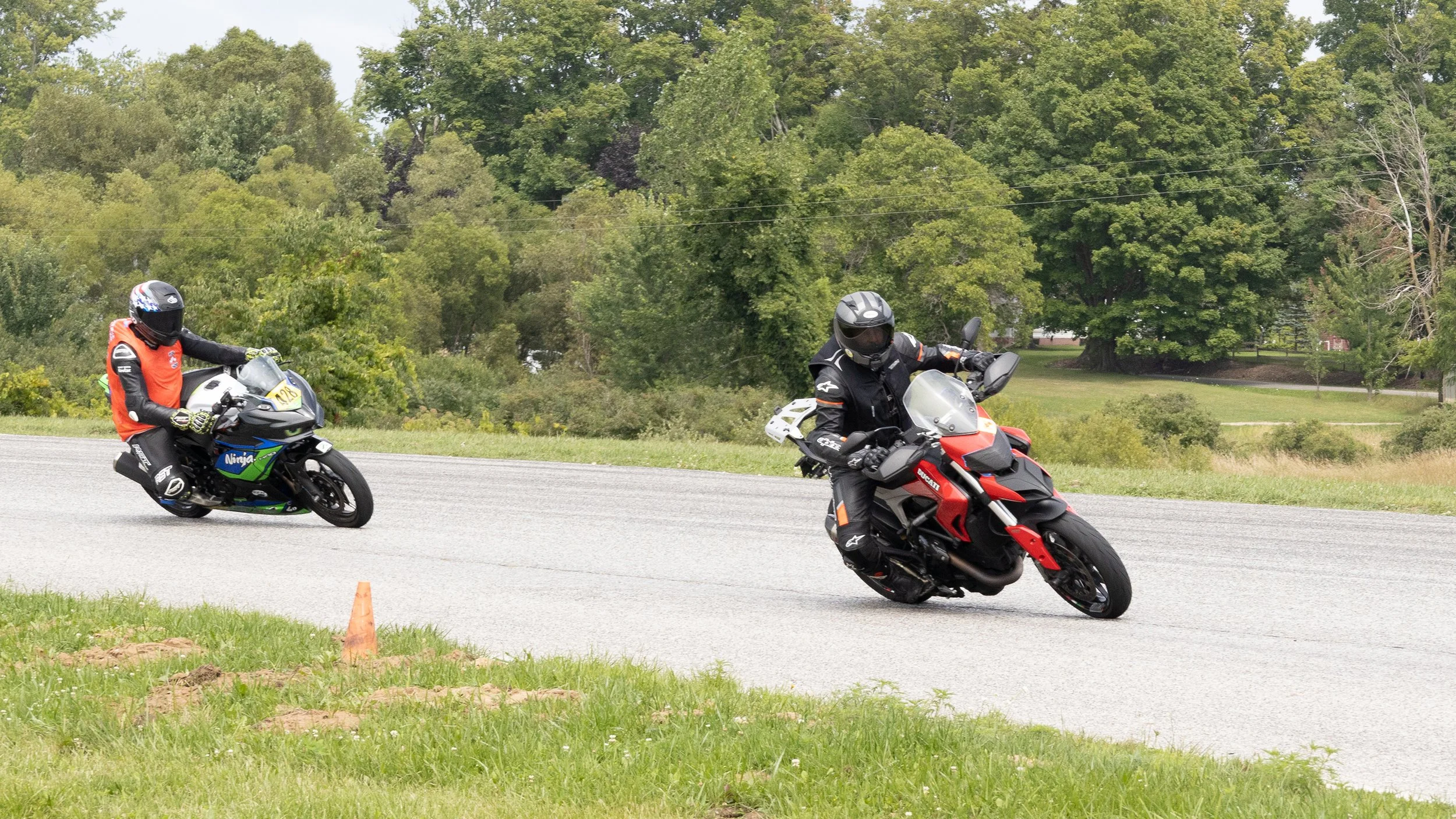 Two motorcyclists riding on a paved course, with one in a black and red motorcycle leaning into a turn, and the other in a black and green motorcycle behind, on a grassy area with trees in the background.