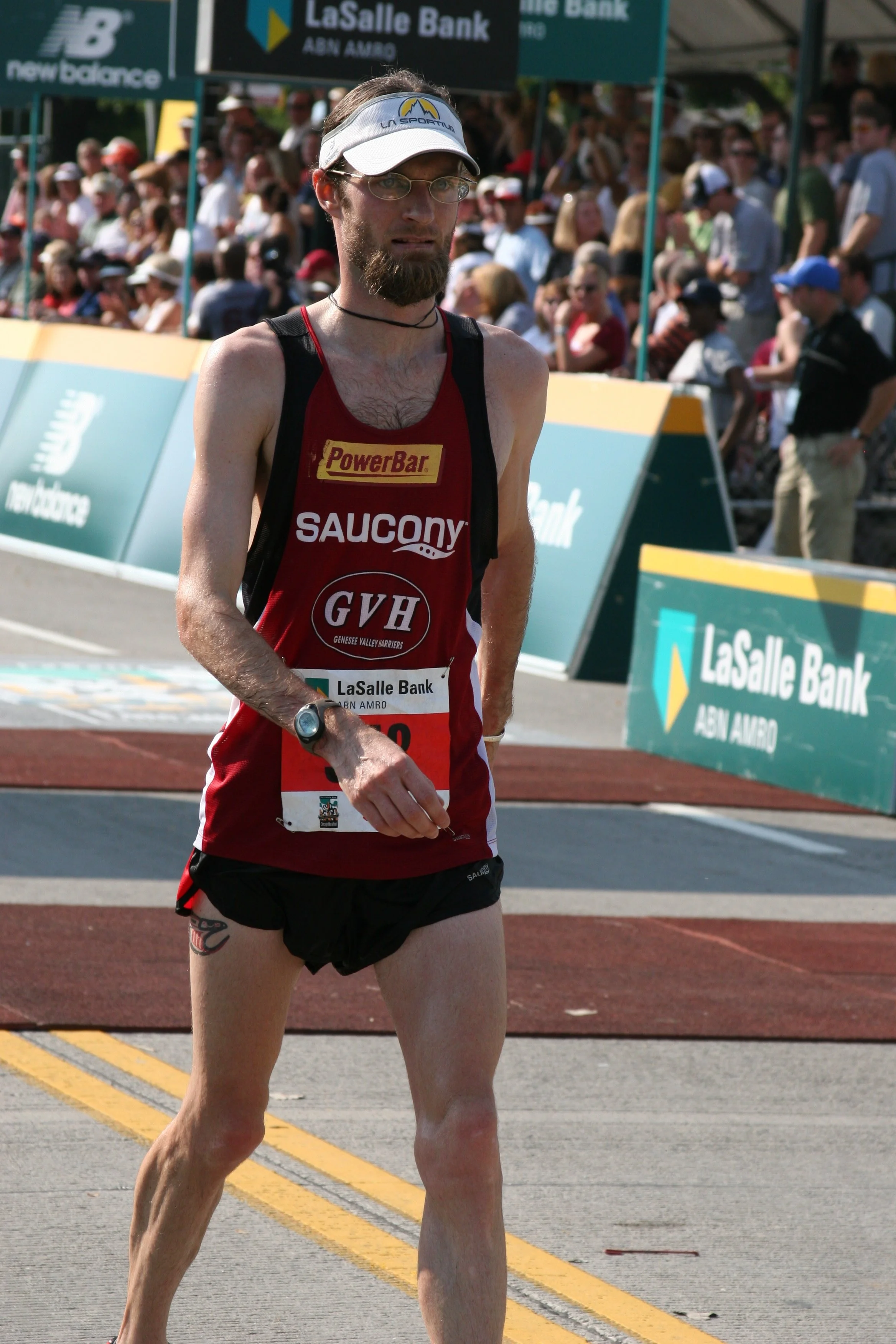 A male marathon runner with a beard, wearing glasses, a white visor, and a red running tank top with sponsor logos, walking near the finish line with a crowd of spectators in the background.
