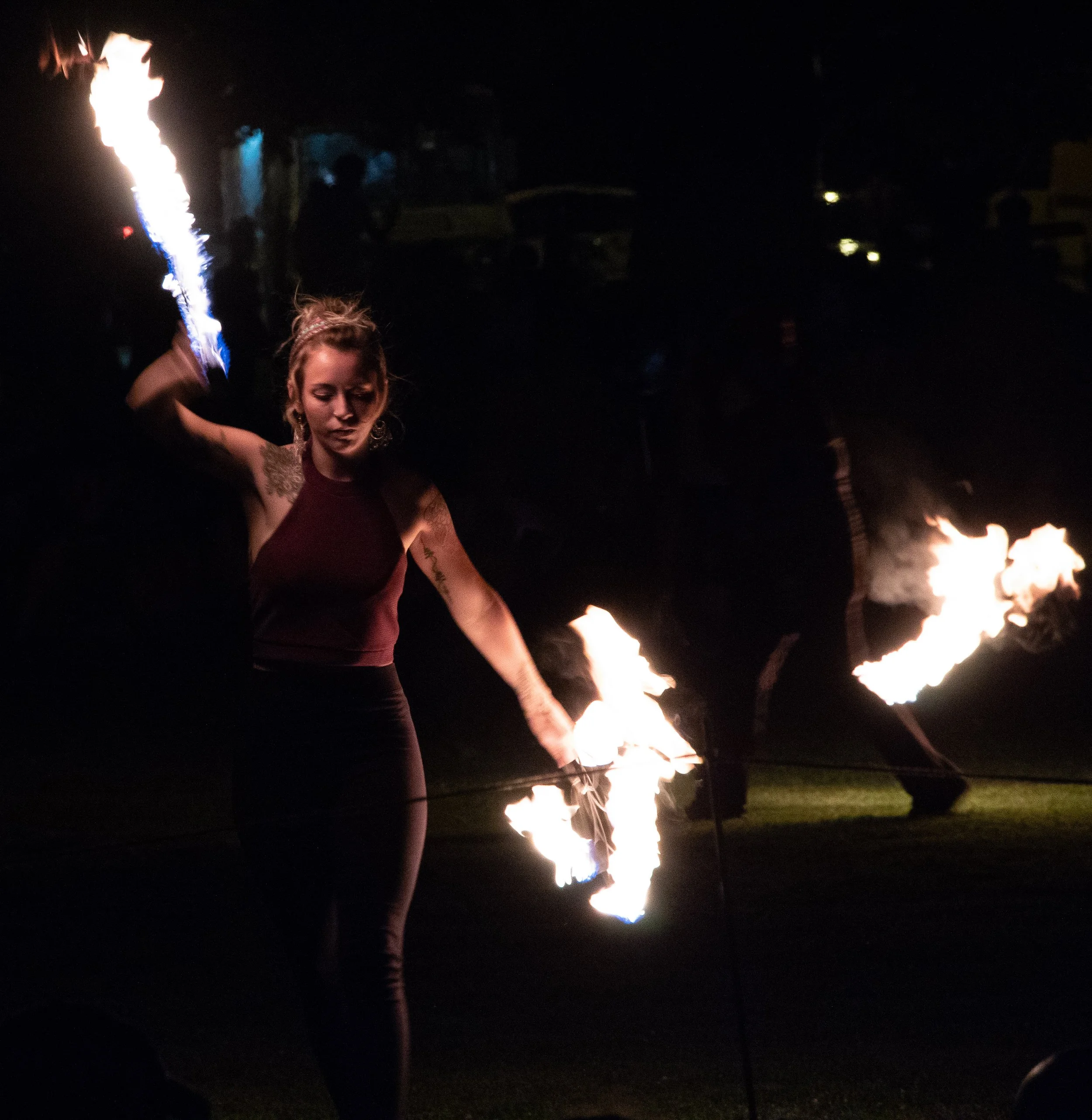 A woman with tattoos on her arms holding a flaming torch at night, with a dark sky and distant lights in the background.