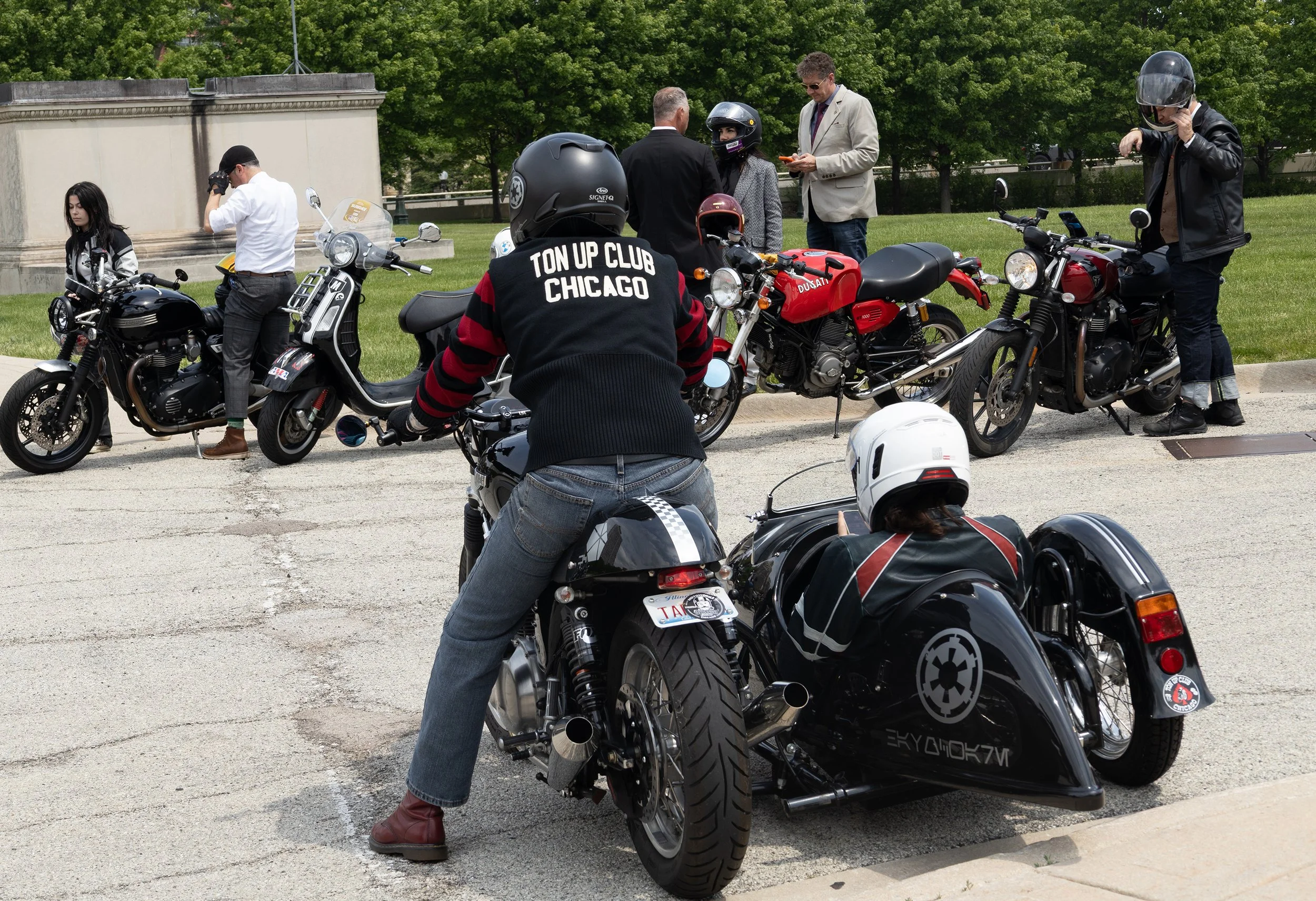 Group of people with motorcycles gathered in a park, some wearing helmets and protective clothing, engaging in conversation.