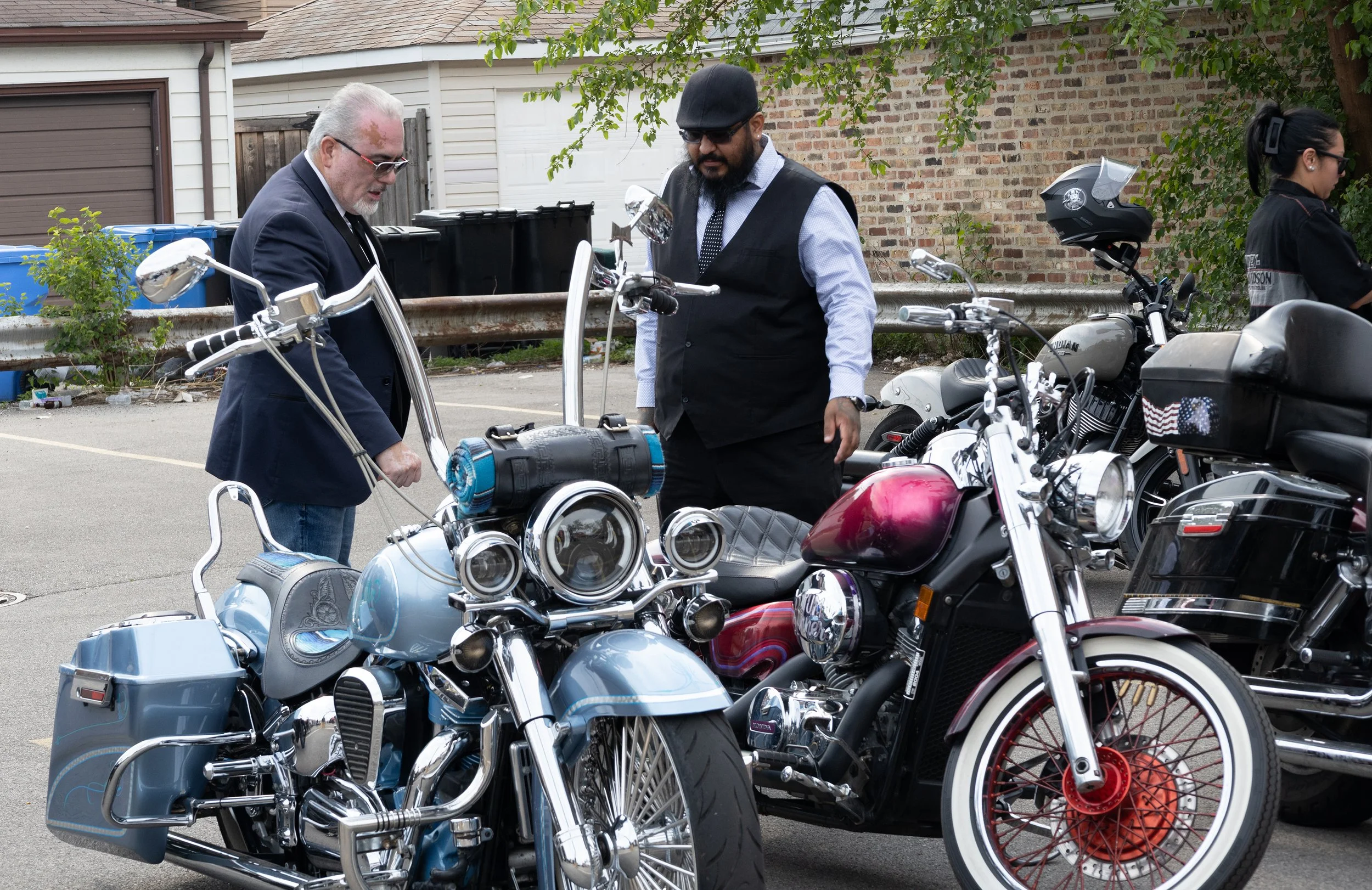 Two men inspecting motorcycles in a parking lot, with one man in a suit and white beard, and another man in a vest with a black hat, black beard, and sunglasses. A third woman with dark hair and sunglasses stands nearby. There are houses and trees in