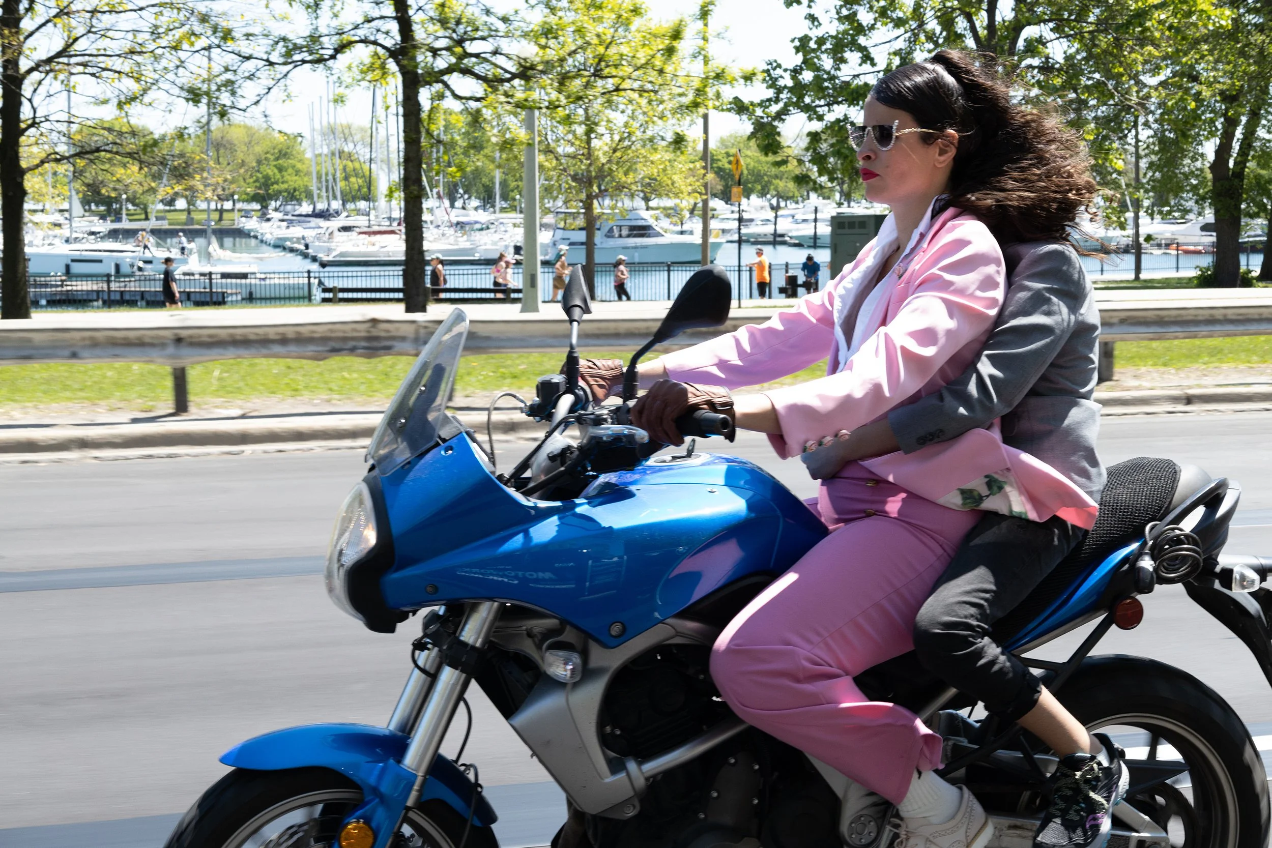 Woman riding a blue scooter along a street with a marina and boats in the background, wearing sunglasses and a pink suit.