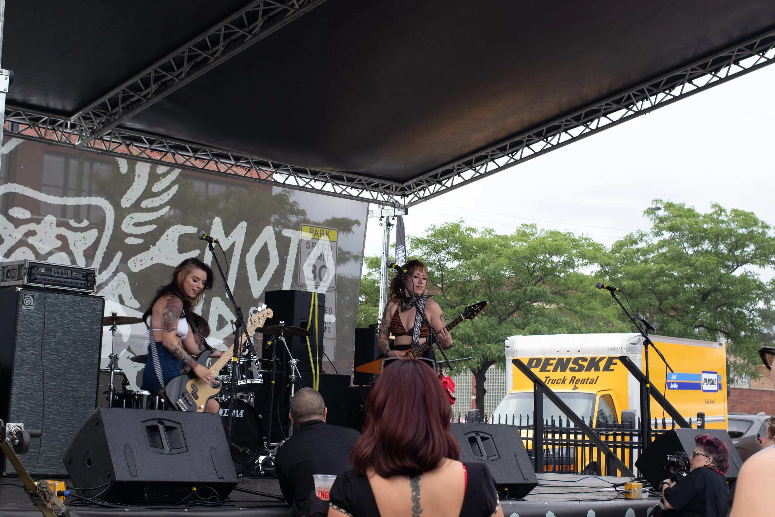 A band performs on an outdoor stage, with musicians playing guitars and singing into microphones. The stage has a black roof, metal framework, and a large backdrop with artwork. Audience members are seated in front, including individuals with tattoos