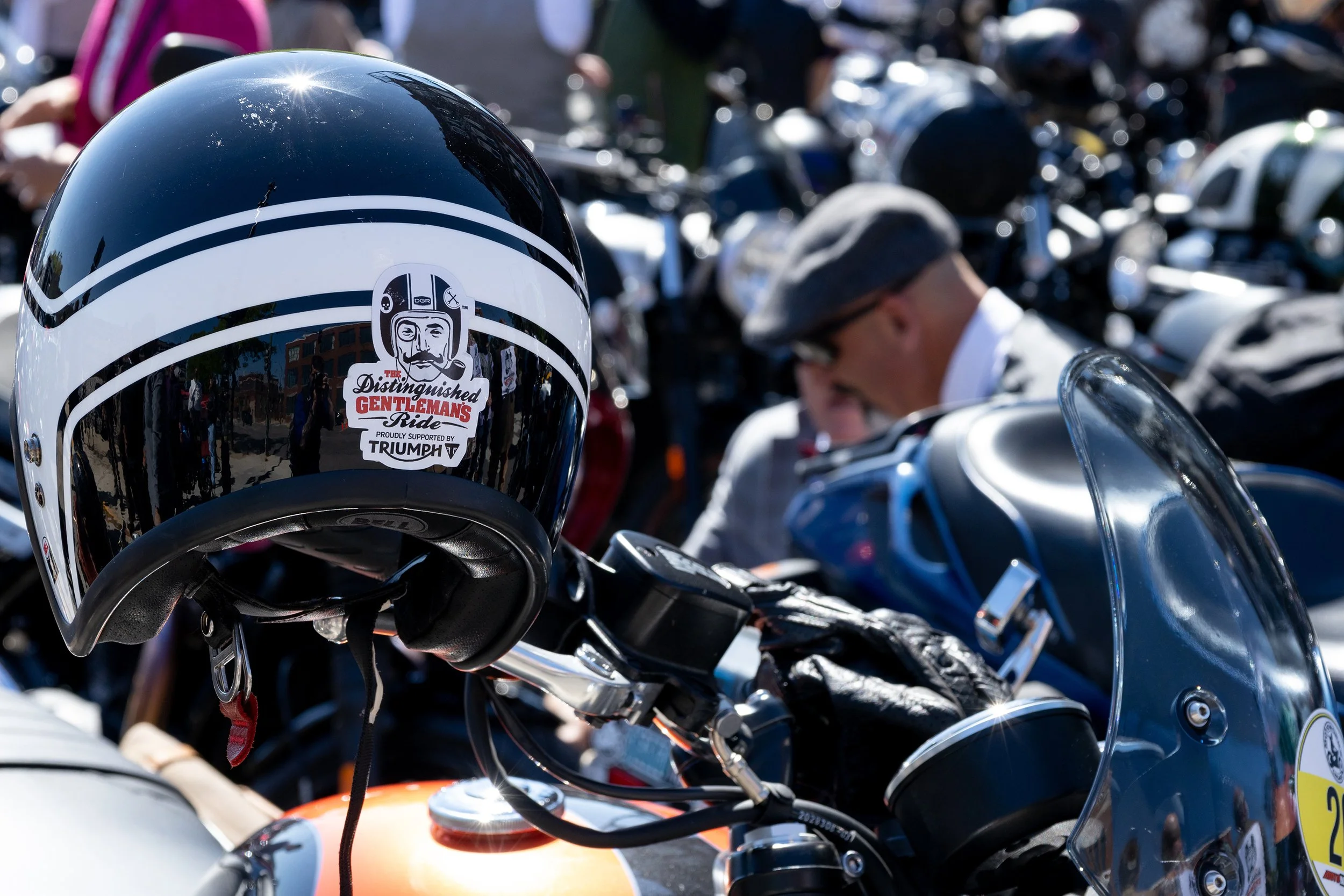 Close-up of a black motorcycle helmet with a sticker reading 'The Distinguished Gentleman's Ride' supported by Triumph in front of a row of motorcycles and people