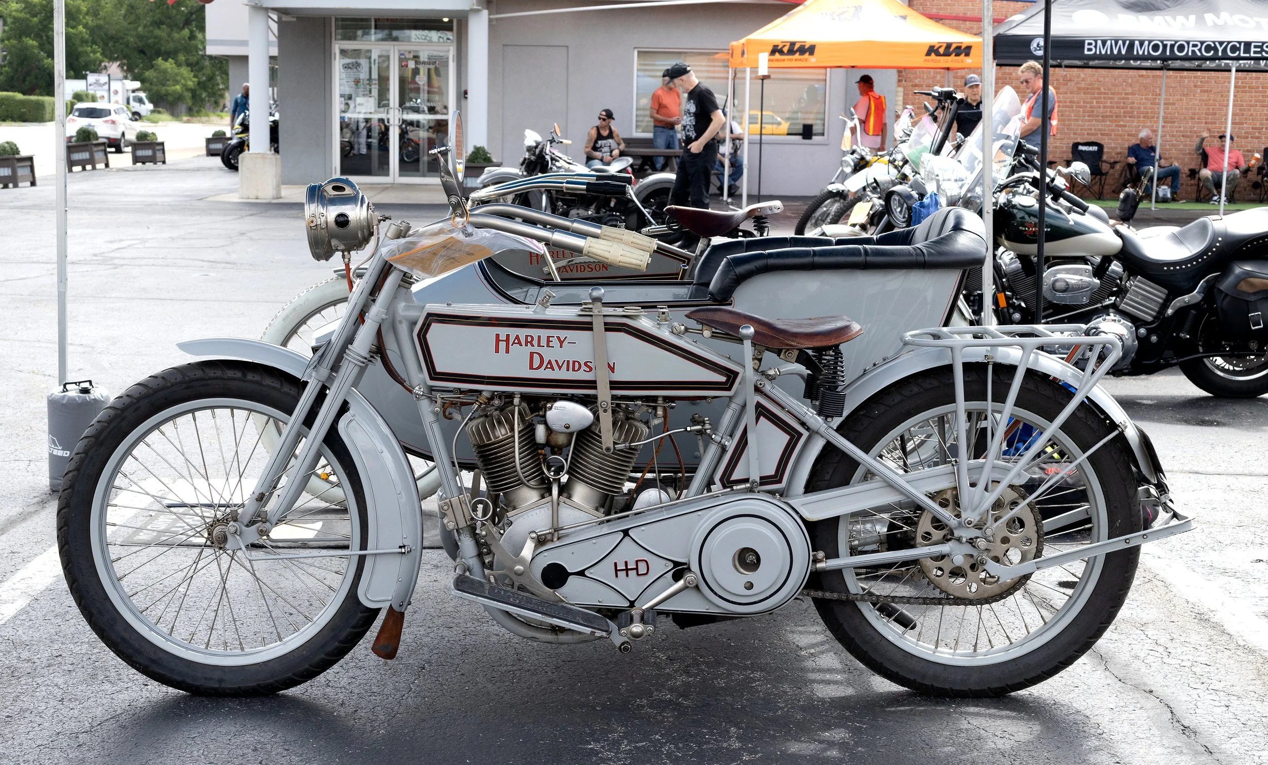 A vintage Harley Davidson motorcycle with a sidecar parked in front of a building, with people and tents in the background at an outdoor event.