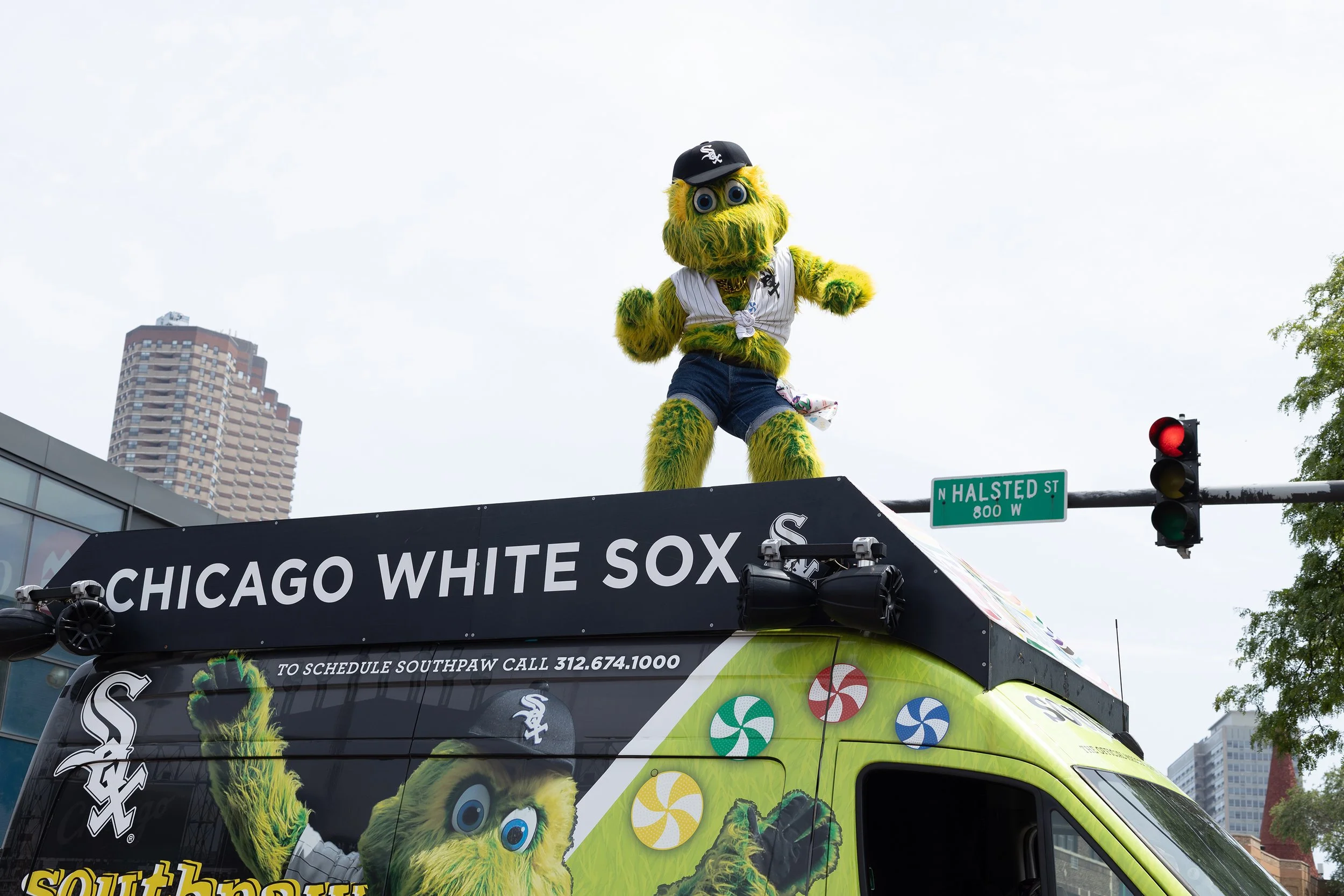 A mascot dressed as the Chicago White Sox baseball team sits on top of a bus with the team's logo and colors, with a cityscape and street sign in the background.