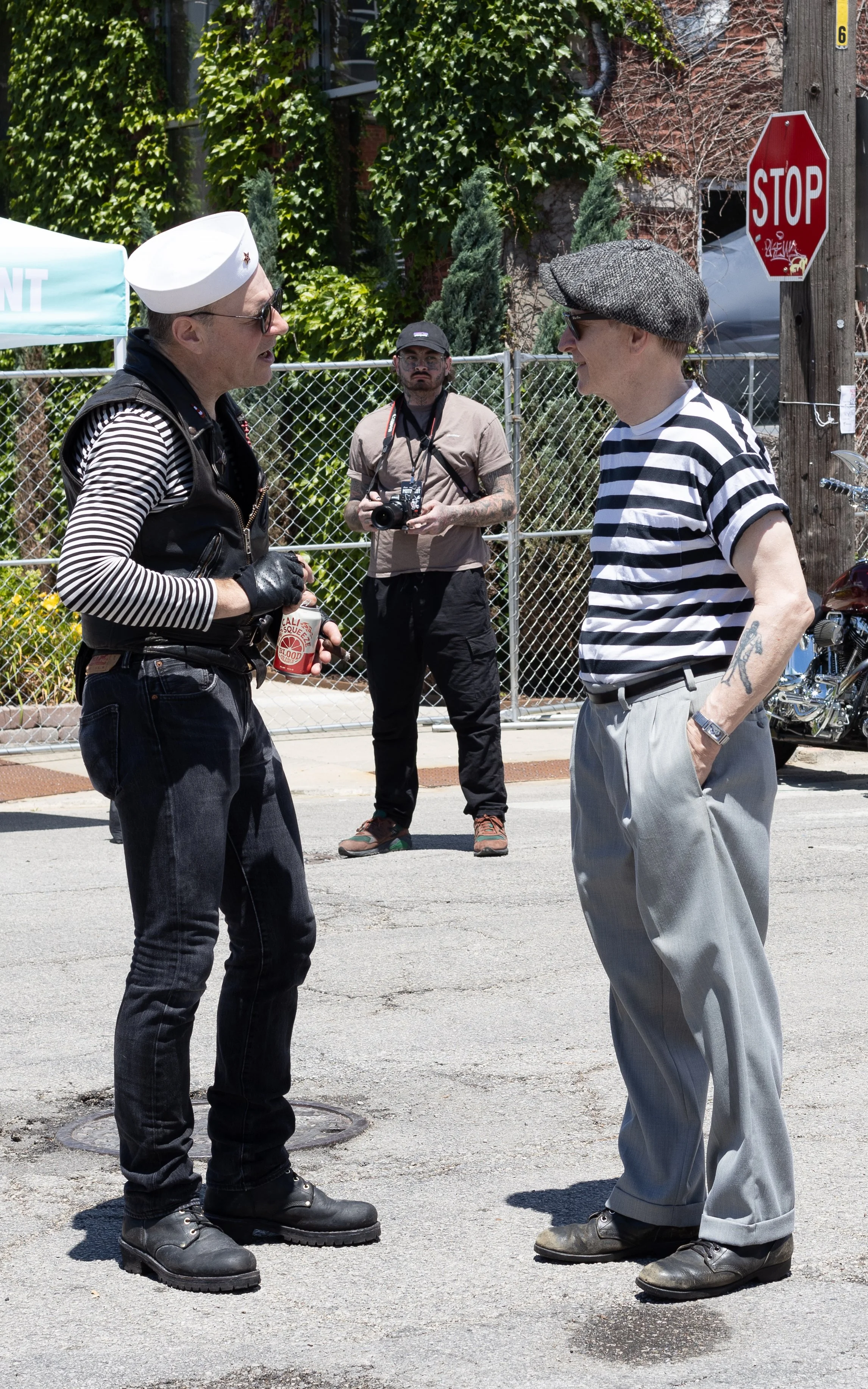 Two men with vintage style clothing stand facing each other on a street, engaged in conversation, with a passerby taking photos in the background.