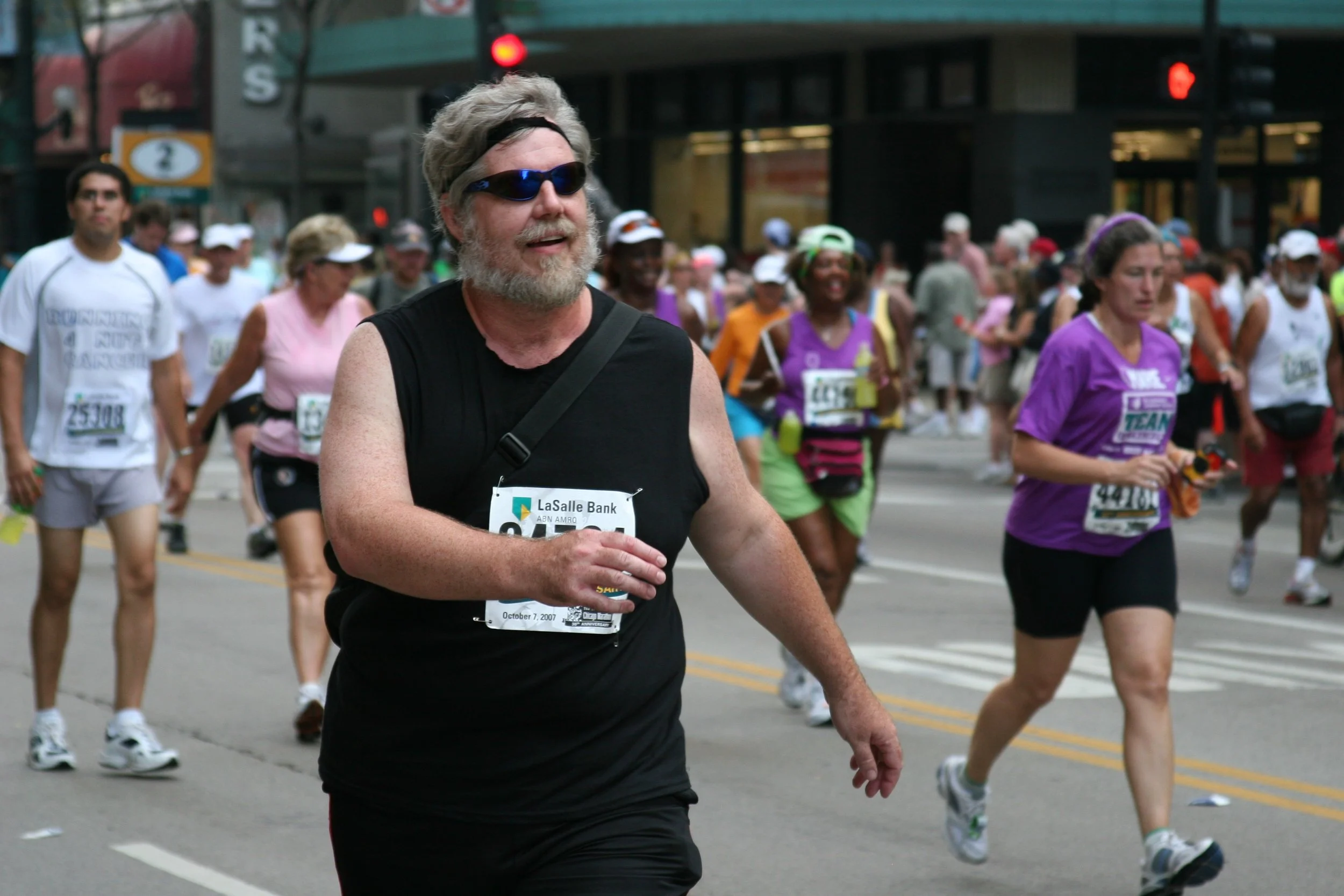 A man with gray hair and beard wearing sunglasses, running in a marathon with a crowd of diverse runners behind him on a city street.