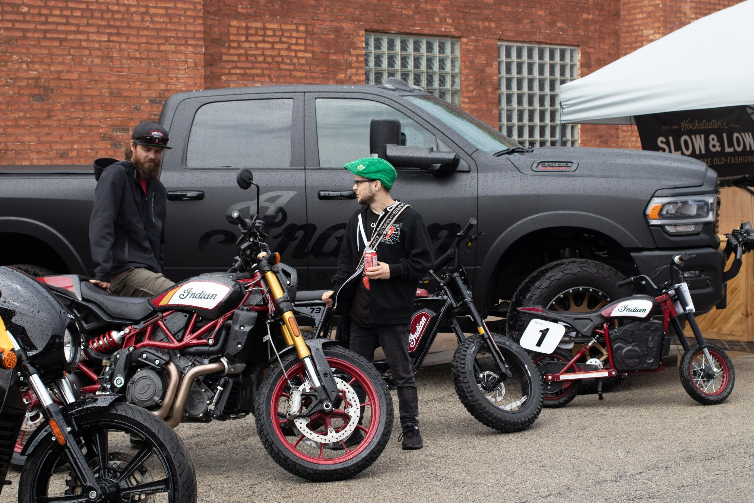 Two men and two Indian motorcycles parked in front of a black pickup truck and brick building, with one man wearing a green cap and holding a soda can, the other with a beard and black jacket, raindrops on the vehicles.