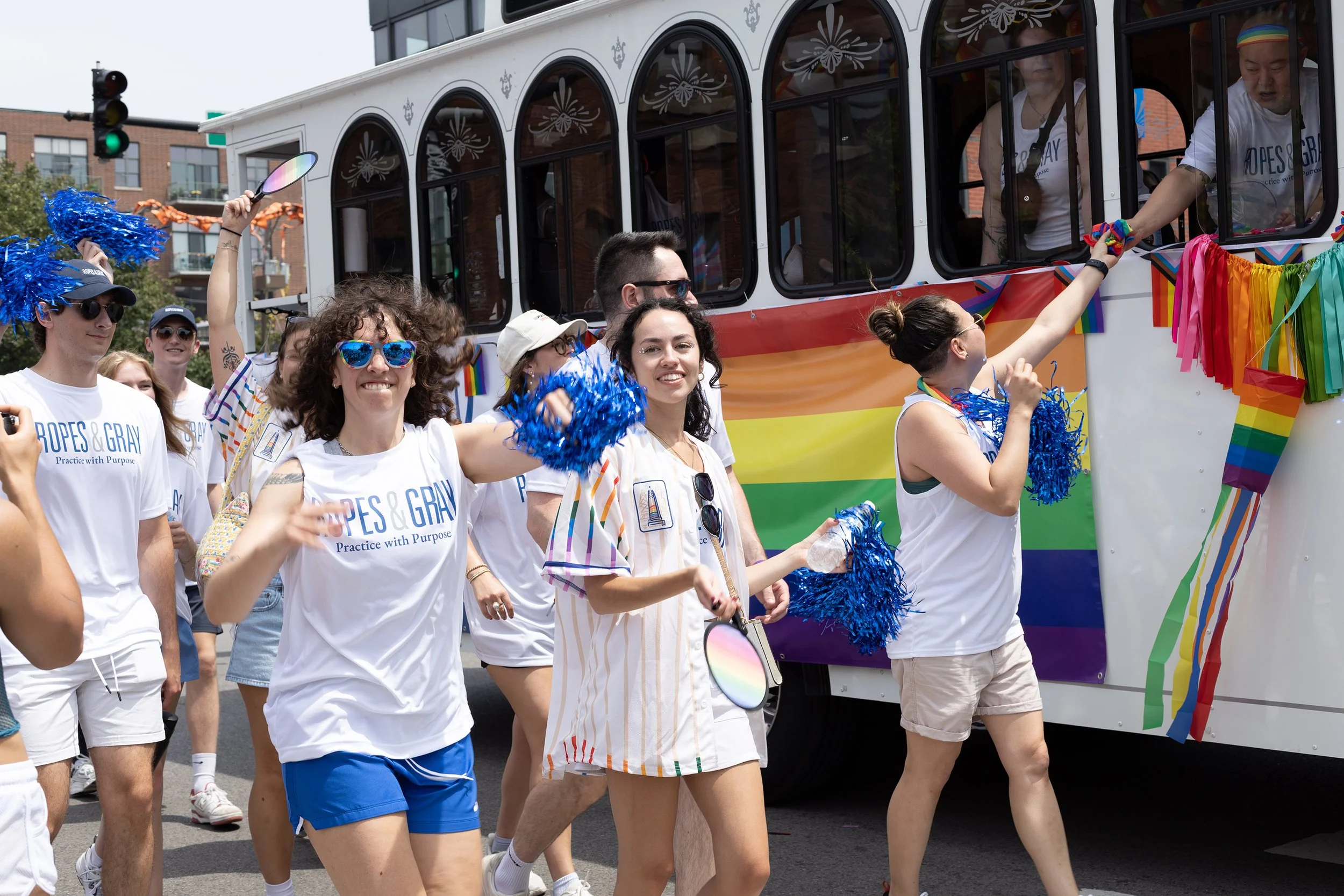 People marching in a pride parade display rainbow decorations and LGBTQ+ flags while walking next to a decorated bus. Some individuals are wearing sunglasses and holding colorful pom-poms, smiling and celebrating.