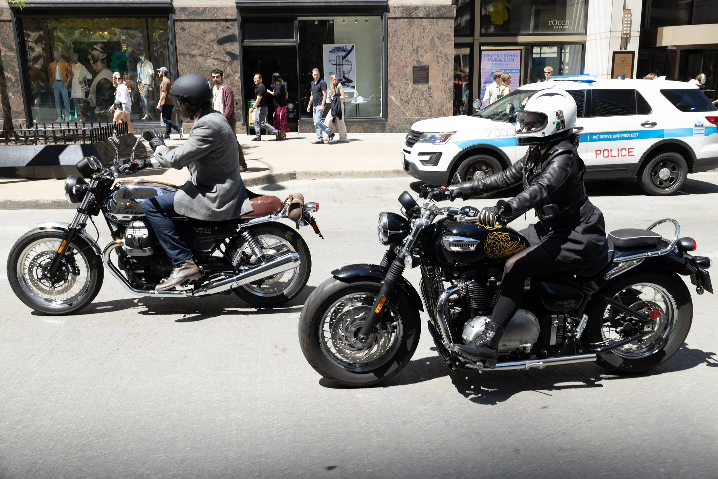 Two motorcyclists, one wearing a gray blazer and the other in black leather, riding motorcycles on a city street near a police car with pedestrians walking past storefronts.