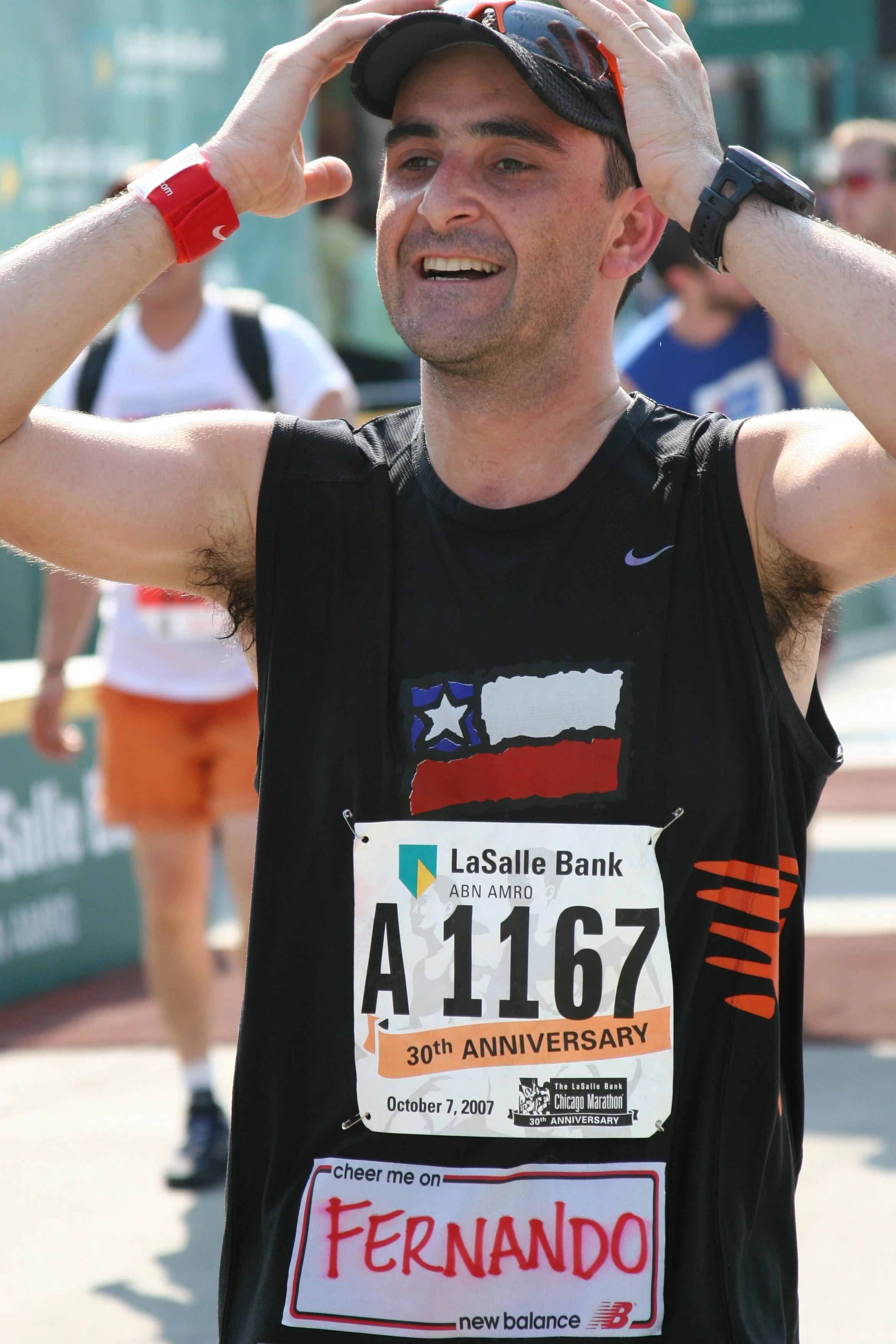 A male marathon runner wearing a black athletic shirt with a race bib from the Chicago Marathon dated October 7, 2007. The bib has the name Fernando and celebrates the 30th anniversary of the race. He is holding his cap while smiling, with sunglasses