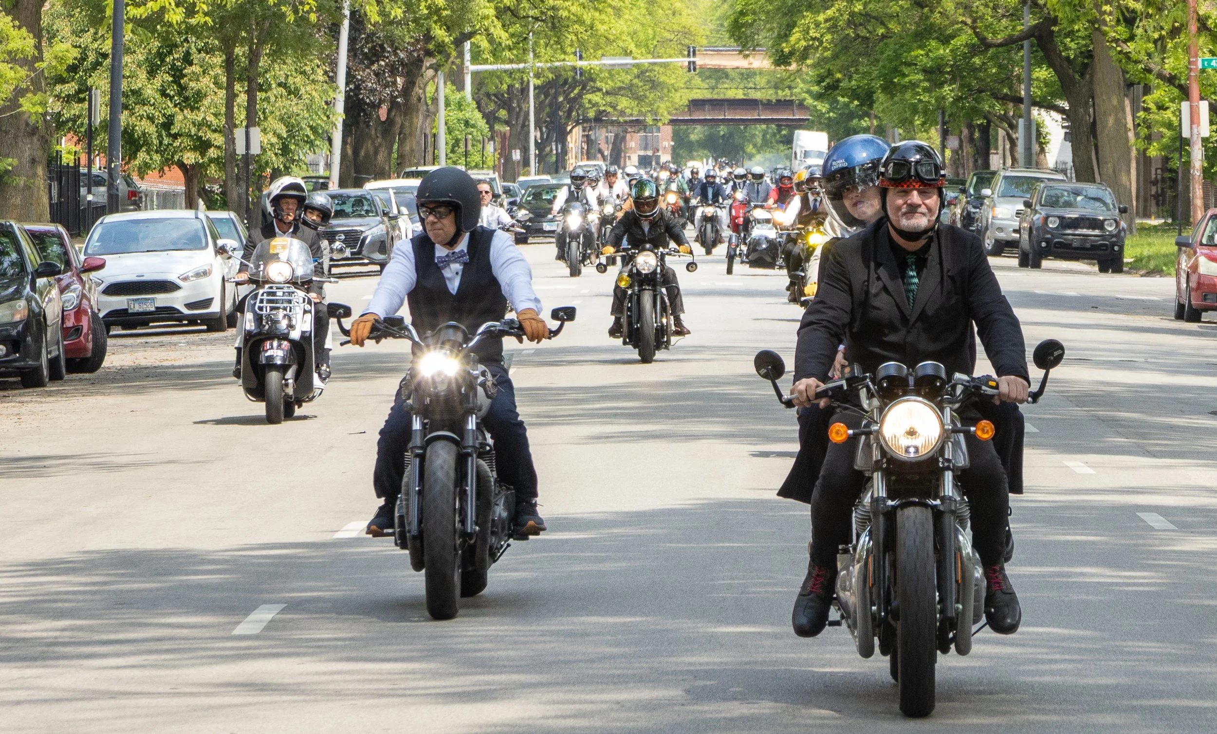 Multiple motorcyclists riding on a city street with cars parked along the sides, green trees lining the road, and a bridge overhead.
