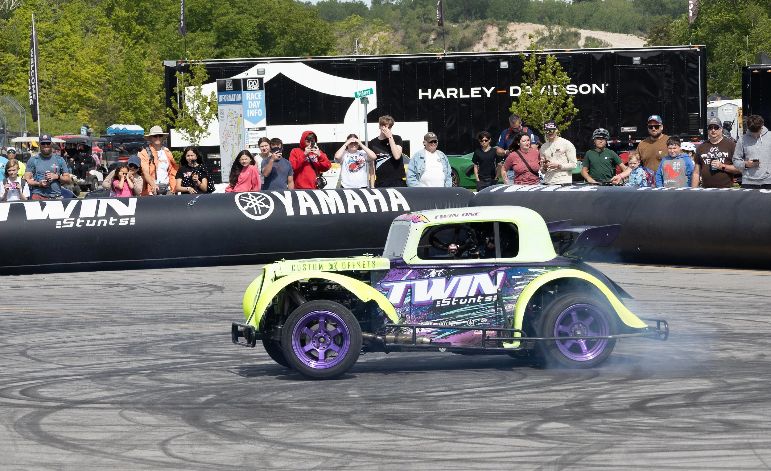 A yellow and purple race car with 'Twins Stunts' written on the side is performing a stunt on a racetrack, creating smoke with its rear tires. Behind the track, spectators are watching, some taking photos or videos, in front of a Harley Davidson trai