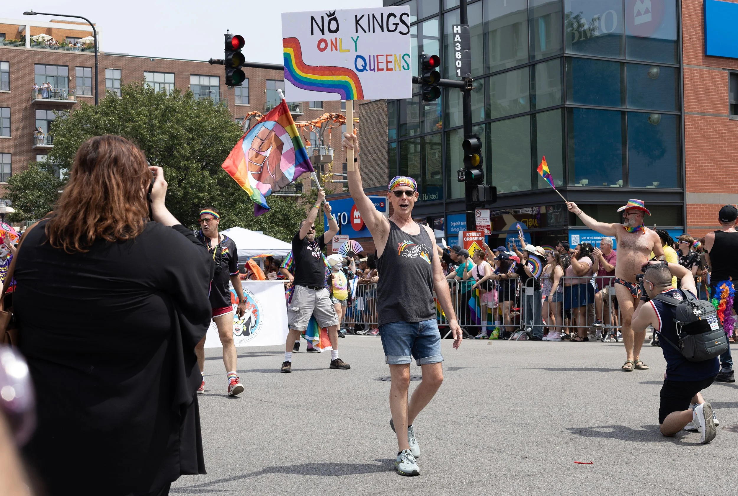 A person at a pride parade holding a sign that says "NO KINGS ONLY QUEENS" with a rainbow-colored rainbow. Other parade participants are visible, including a person with a rainbow flag, rainbow accessories, and showing off pride flags. The scene take