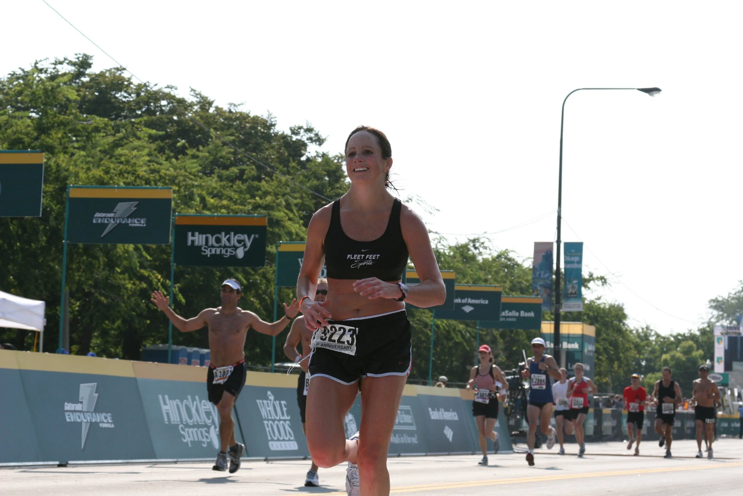 A group of runners participating in a race on a city street, with a woman in the forefront wearing a black tank top and running shorts, smiling as she runs. Key banners advertising the event are visible, and a background of trees and street signs alo