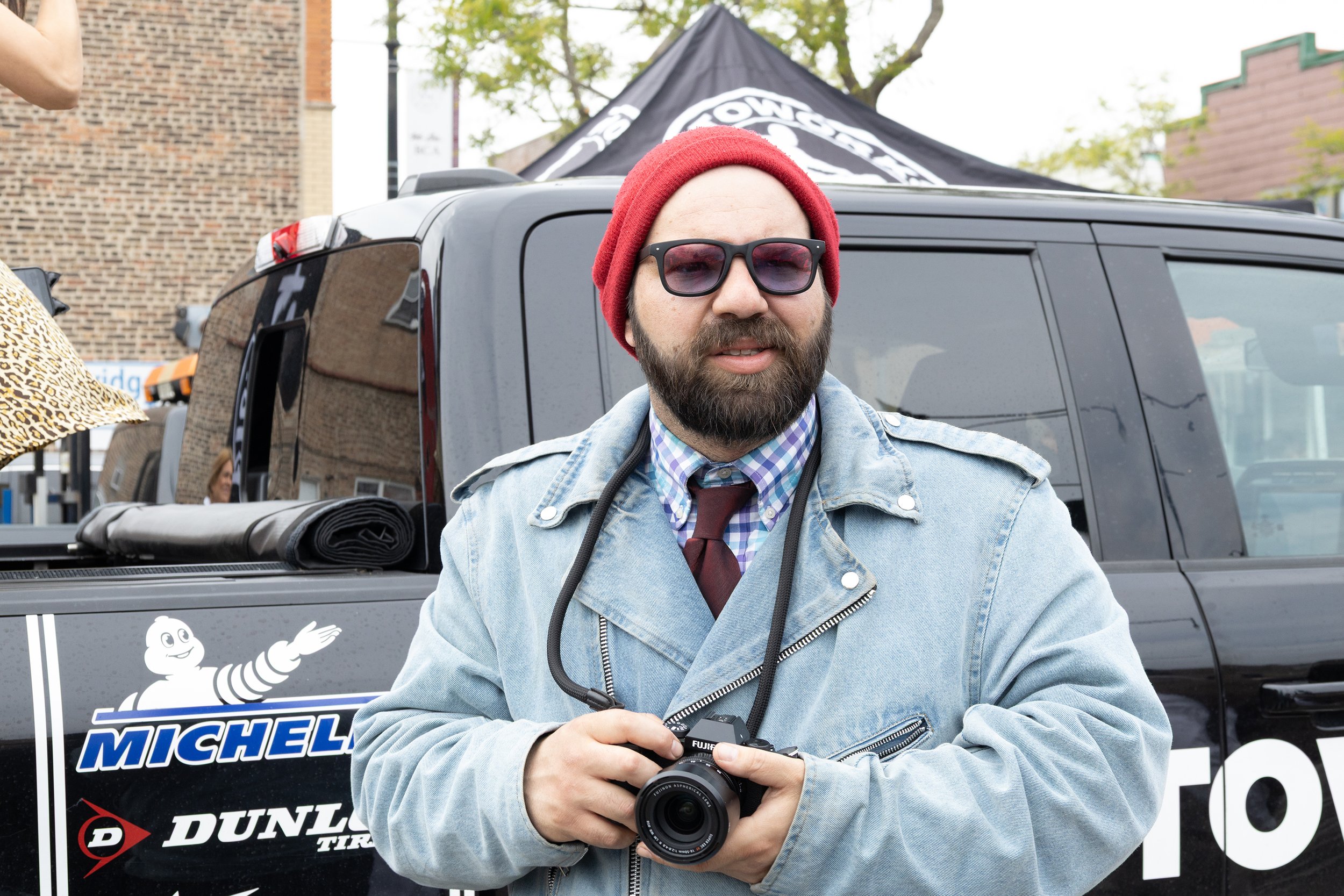 A bearded man wearing sunglasses, a red beanie, a denim jacket, and a checkered shirt, holding a camera, standing in front of a black SUV.