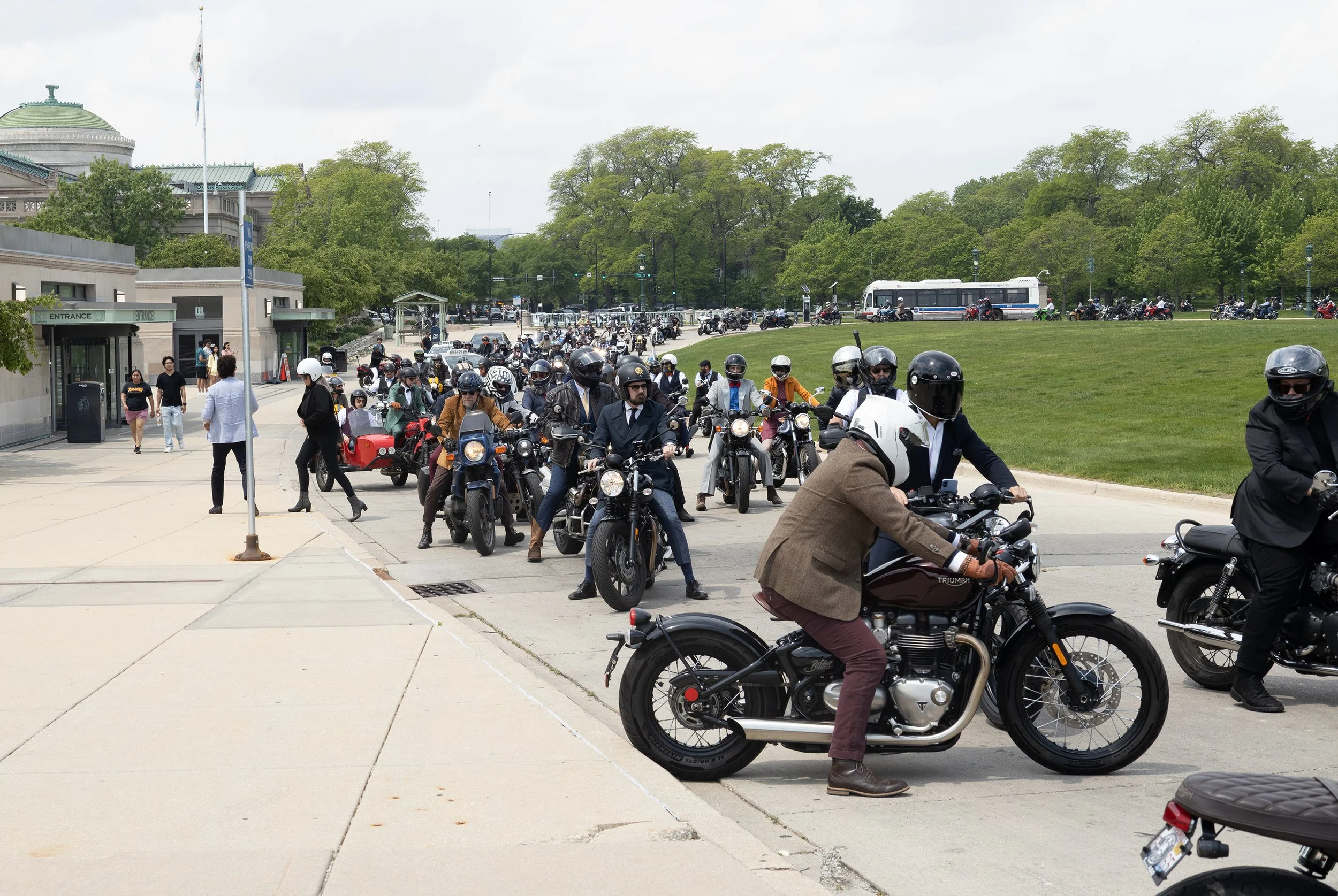 A large group of motorcyclists and scooters gathered and riding on a city street near a park, with green trees and historic buildings in the background.