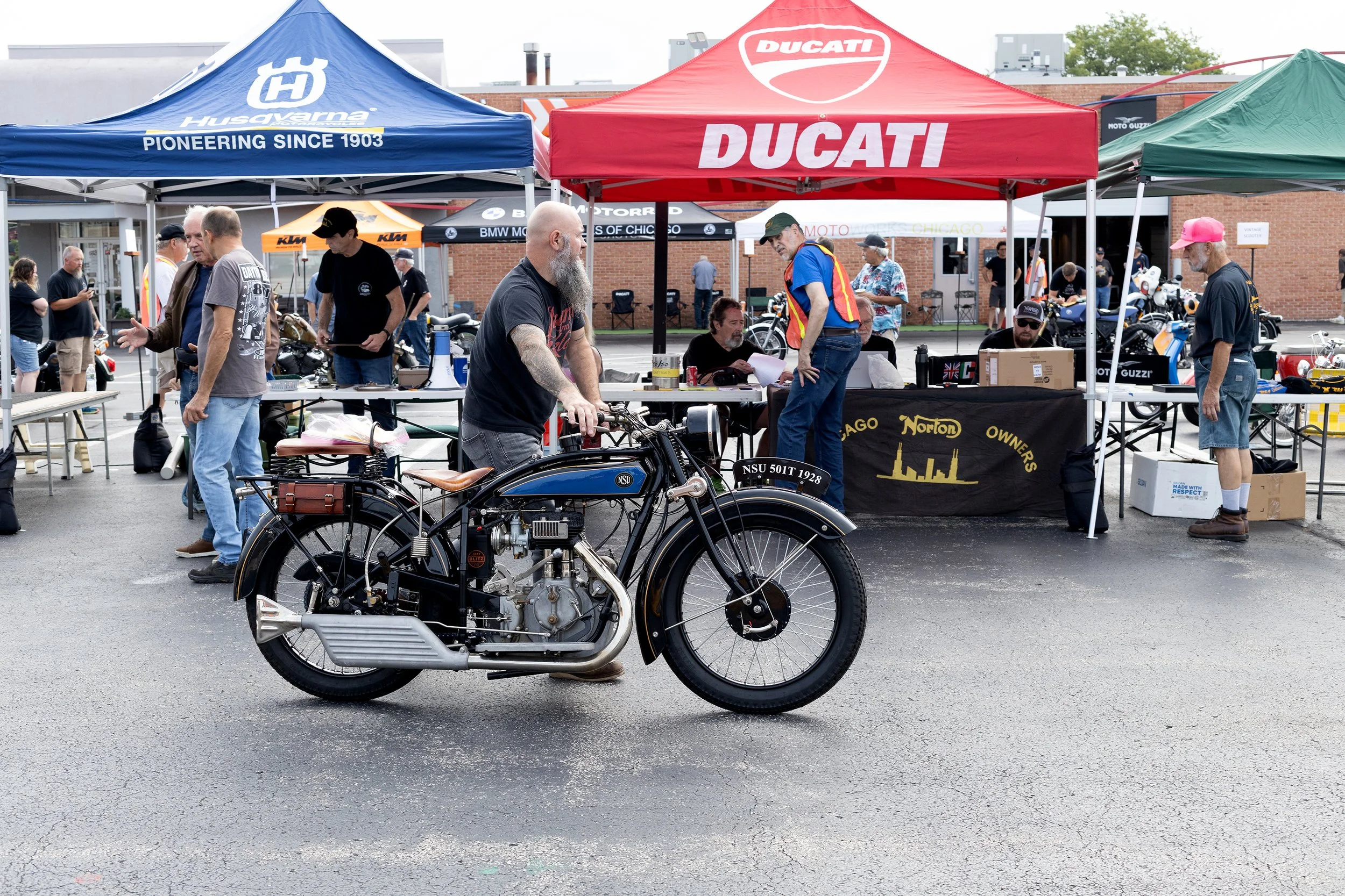 A vintage motorcycle parked in front of tents at an outdoor event with people walking and talking nearby.