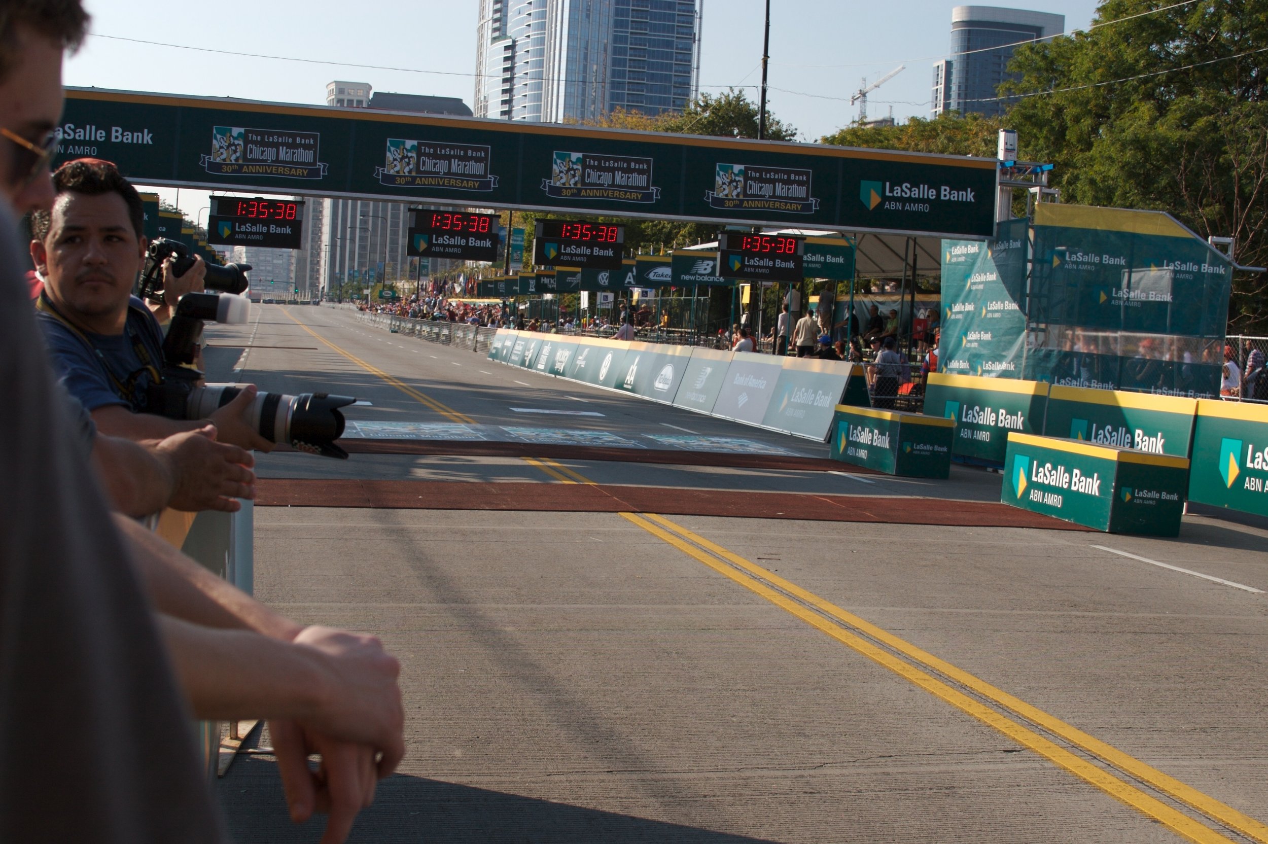Street scene with digital timers showing 1:35:38 at the finish line of a race, with banners for the Chicago Marathon and LaSalle Bank in the background and spectators on the sidewalk.