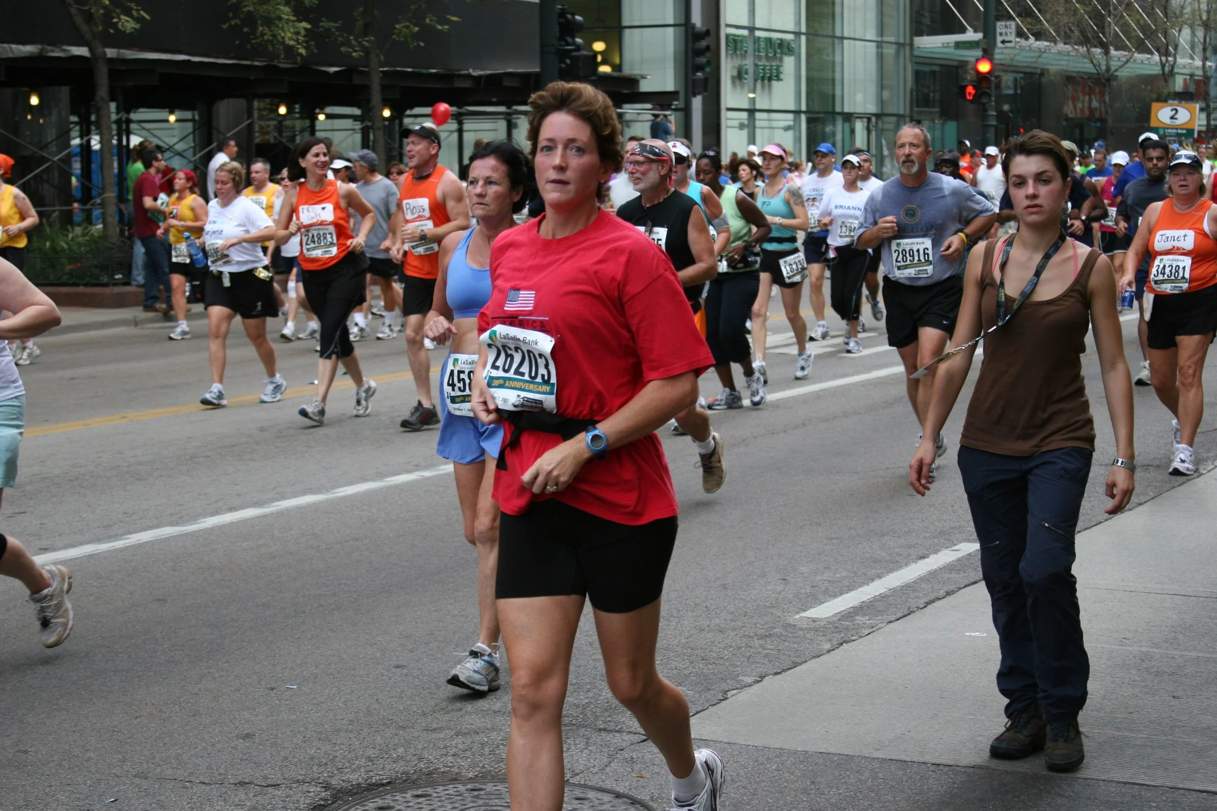 A diverse group of people participating in a marathon or running event on an urban city street, with some wearing race bibs and athletic clothing.