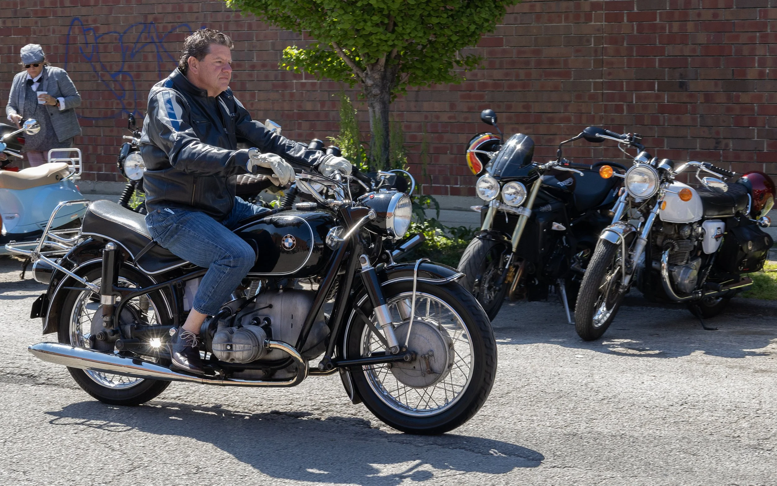 A man riding a black BMW motorcycle on a street with parked motorcycles against a brick wall and a woman standing nearby in the background.