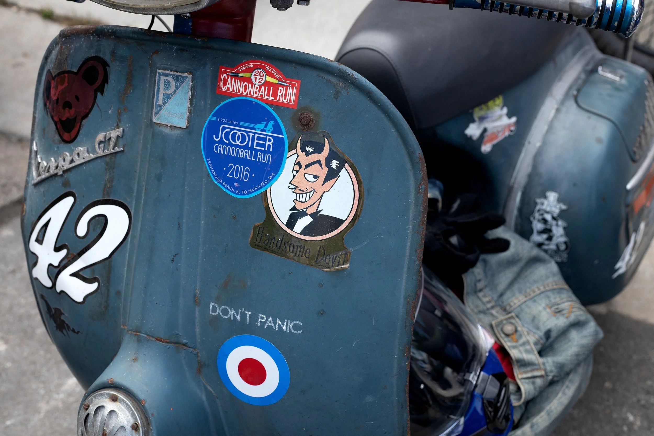 Close-up of a vintage blue scooter decorated with various stickers, including one for a Cannonball Run event with a cartoon devil, and a target symbol, parked on a rough pavement surface.