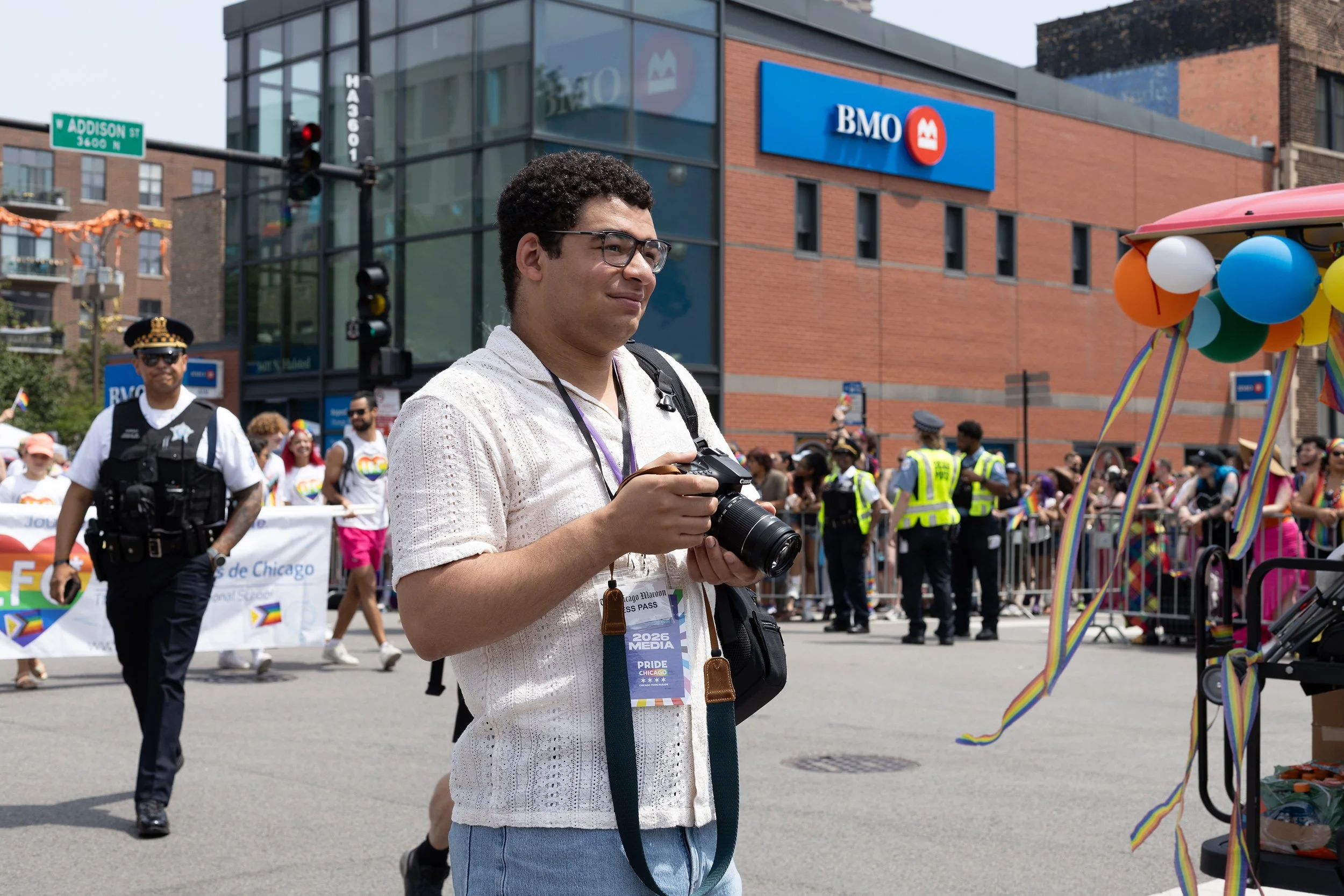 A man with glasses holding a camera at a Pride parade in Chicago, with other parade participants and police officers in the background, and colorful balloons on the right