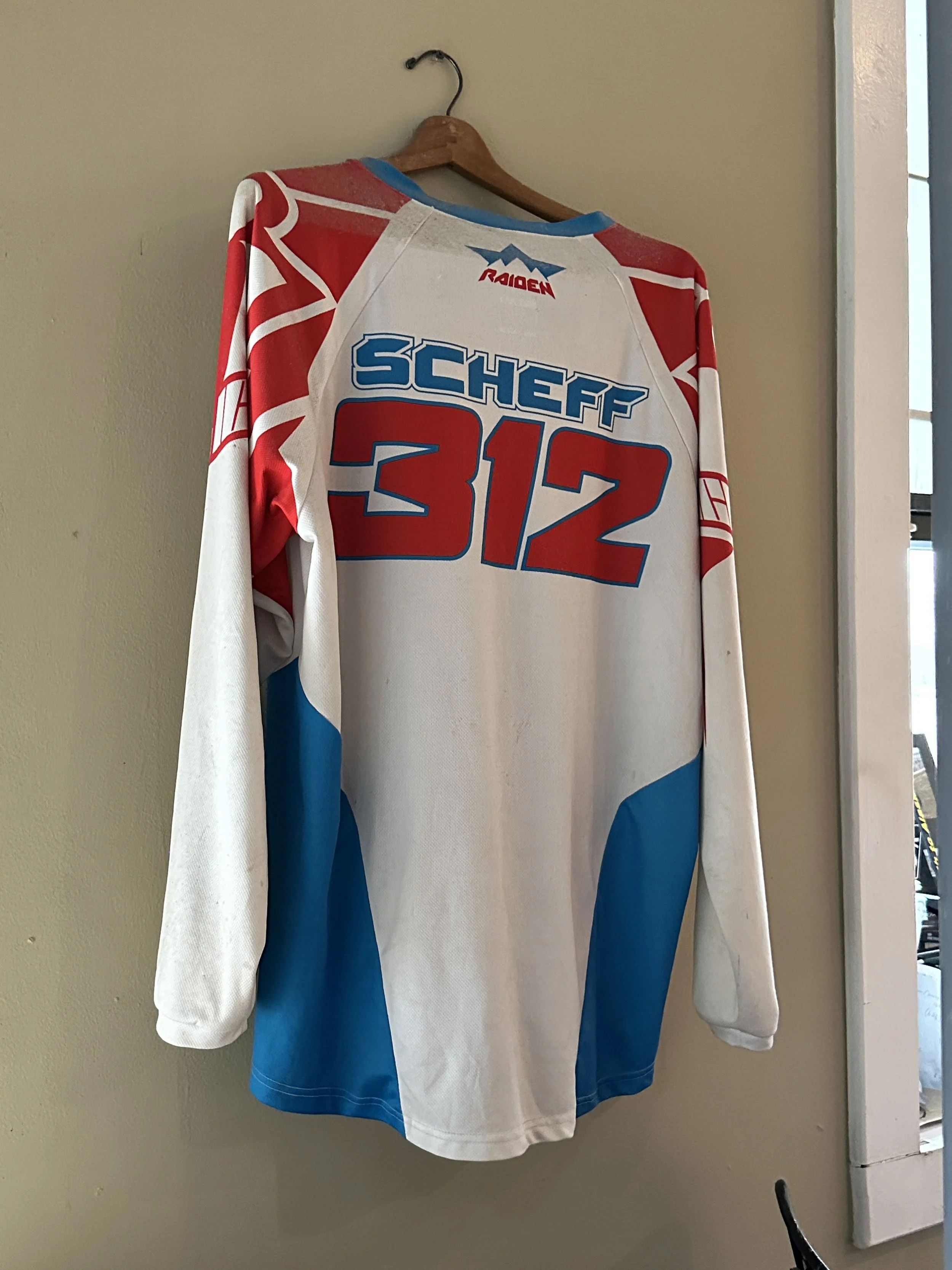 A hockey jersey hanging on a wooden hanger on a wall. The jersey has red, white, and blue colors with the number 312 on the back and the name Scheff. There are some logos on the jersey, including Raiden.