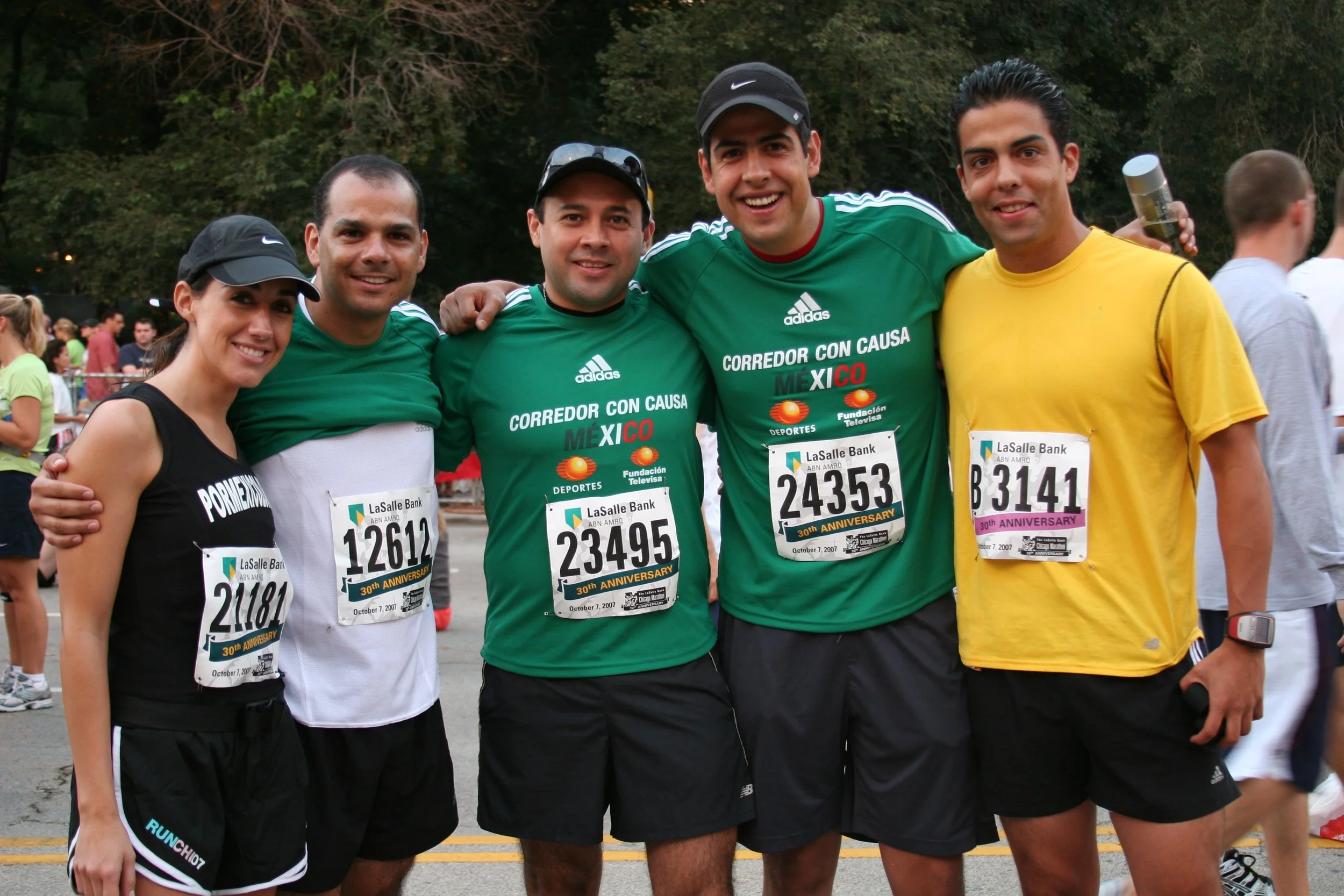 A group of five runners celebrating after a race, standing close together with arms around each other's shoulders, smiling at the camera, at a park or event area with trees and other people in the background.