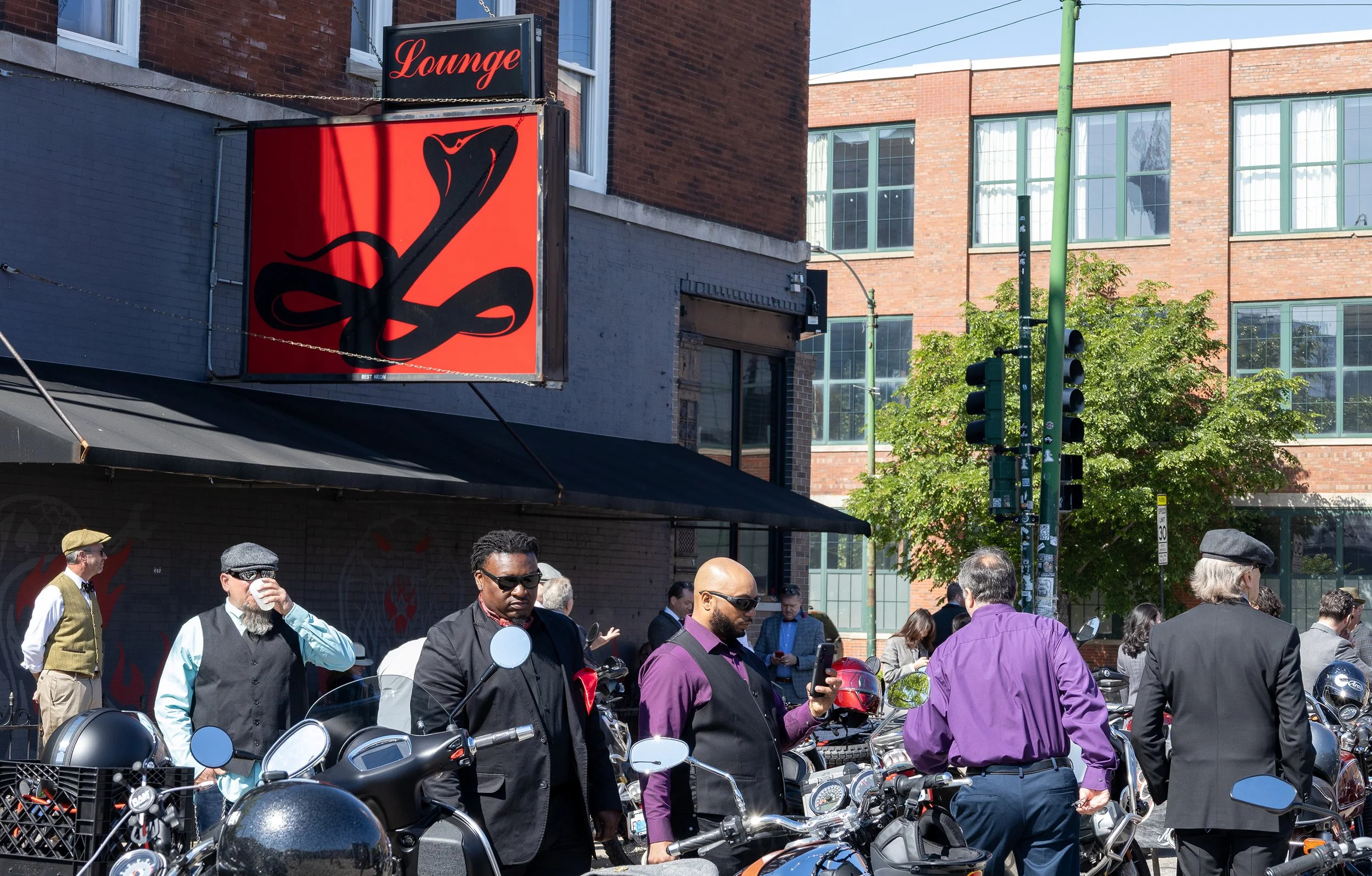 People gathered around scooters on a city street with a red billboard sign reading 'Lounge' and a silhouette of a snake, brick buildings, and green traffic lights in the background.