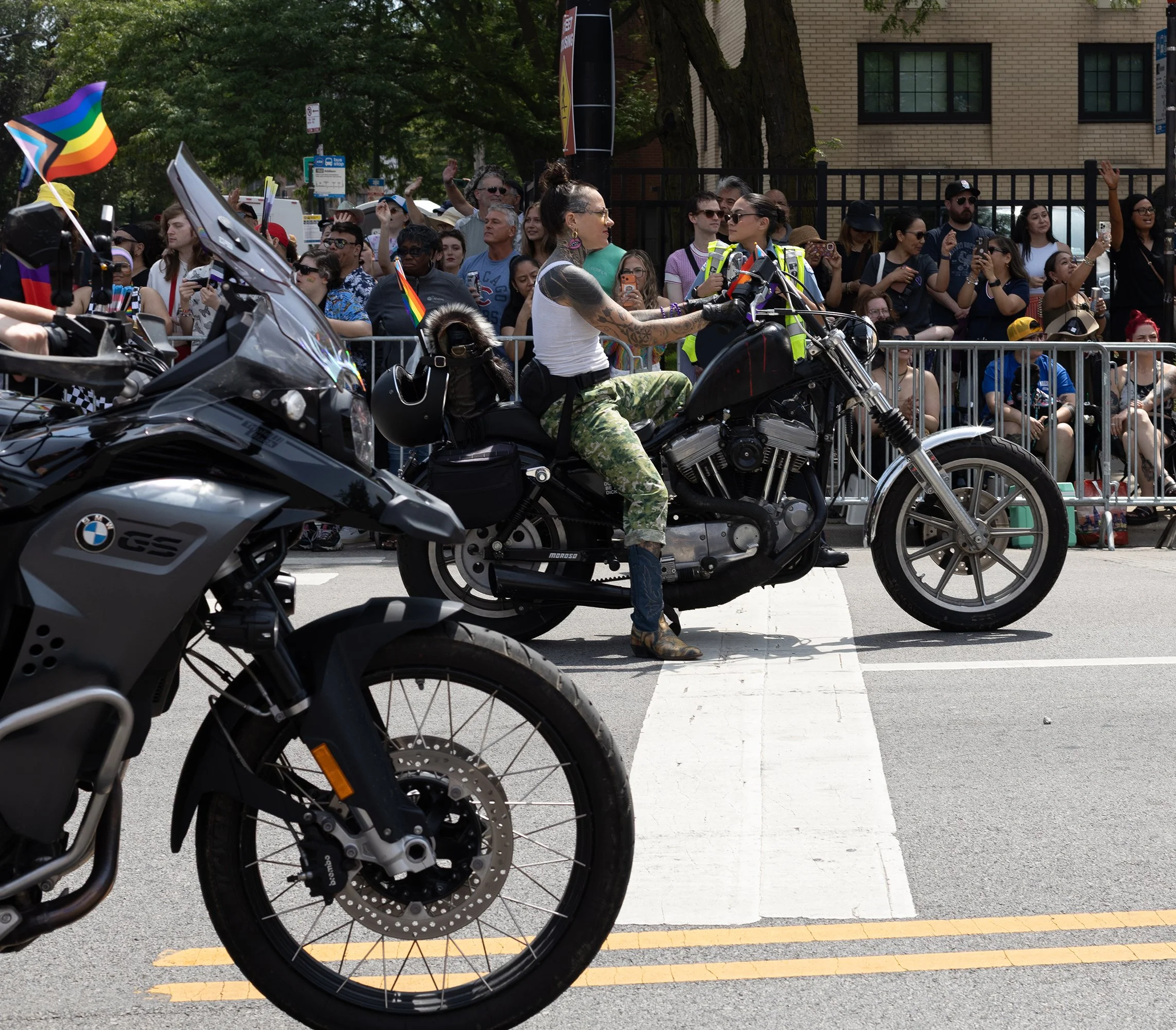 A woman riding a black motorcycle on a street during a parade or event, with a crowd of people, some holding pride flags, gathered behind a metal barrier.