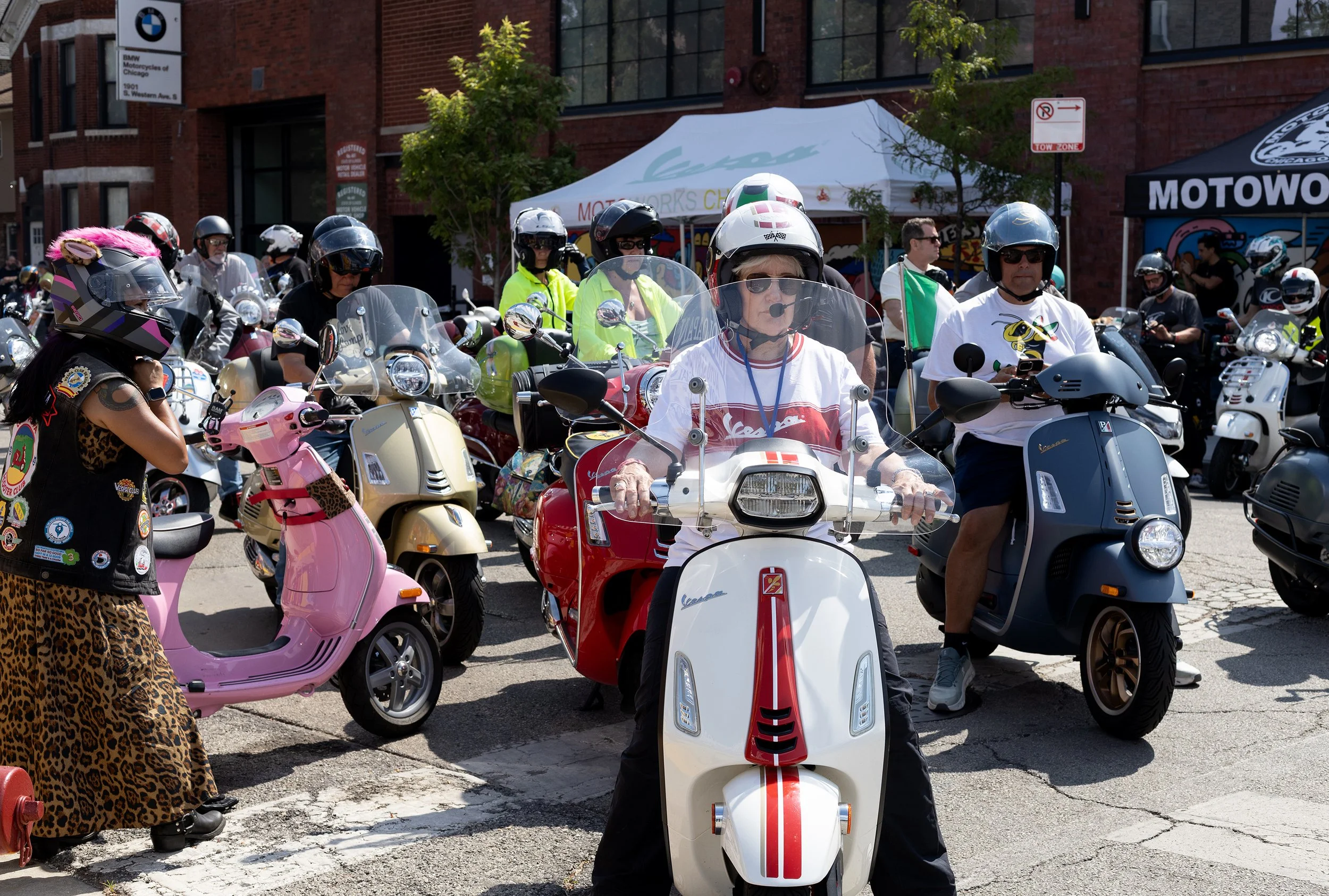 Group of people on scooters gathered outdoors in front of a brick building with a BMW motorcycles sign, some wearing helmets and sunglasses, some with reflective safety vests, with a white tent in the background.