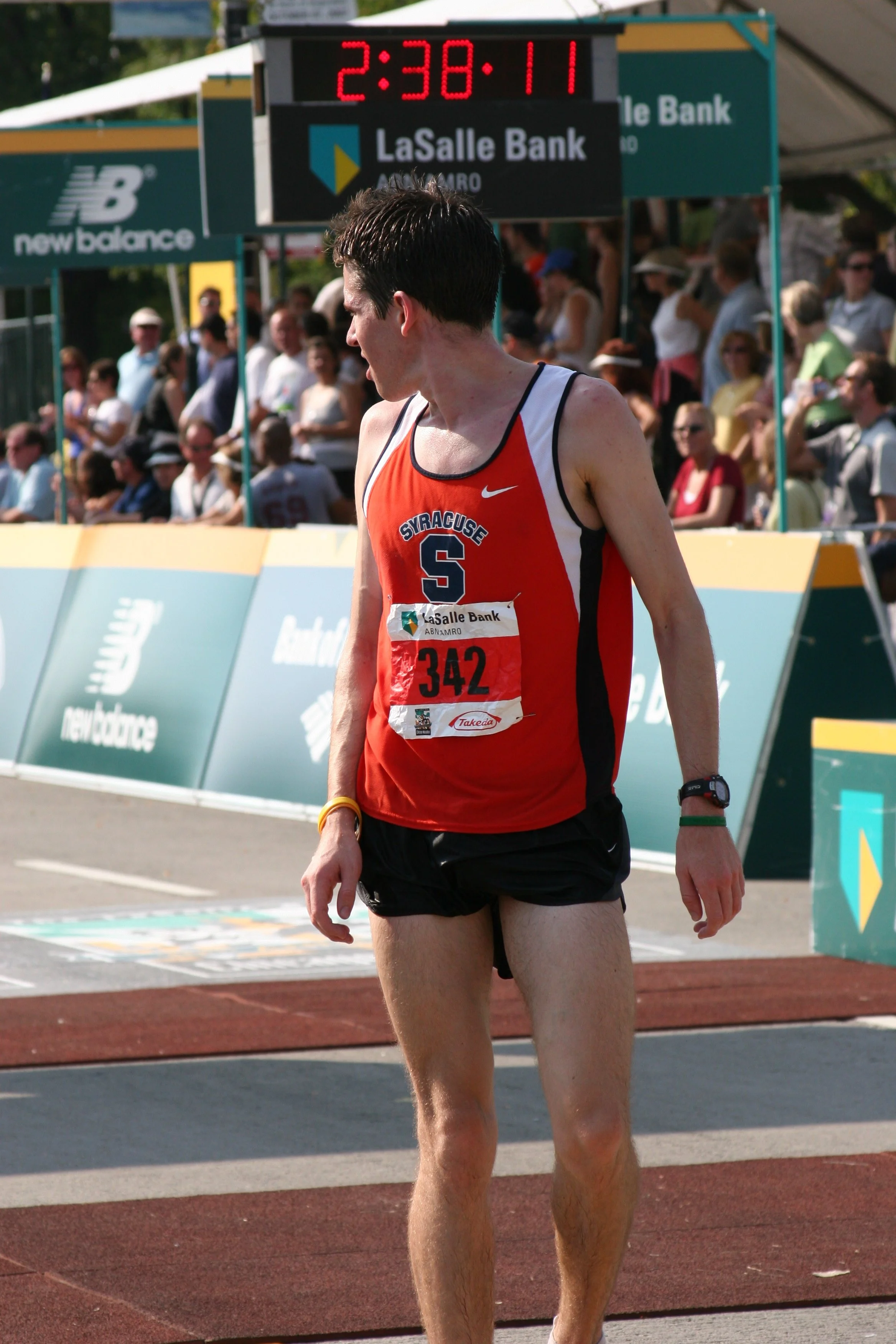 A male runner in a red and black Syracuse University shirt and black shorts, near the finish line of an outdoor race, with a large clock showing 2:38:11 in the background and a crowd of spectators behind barriers.