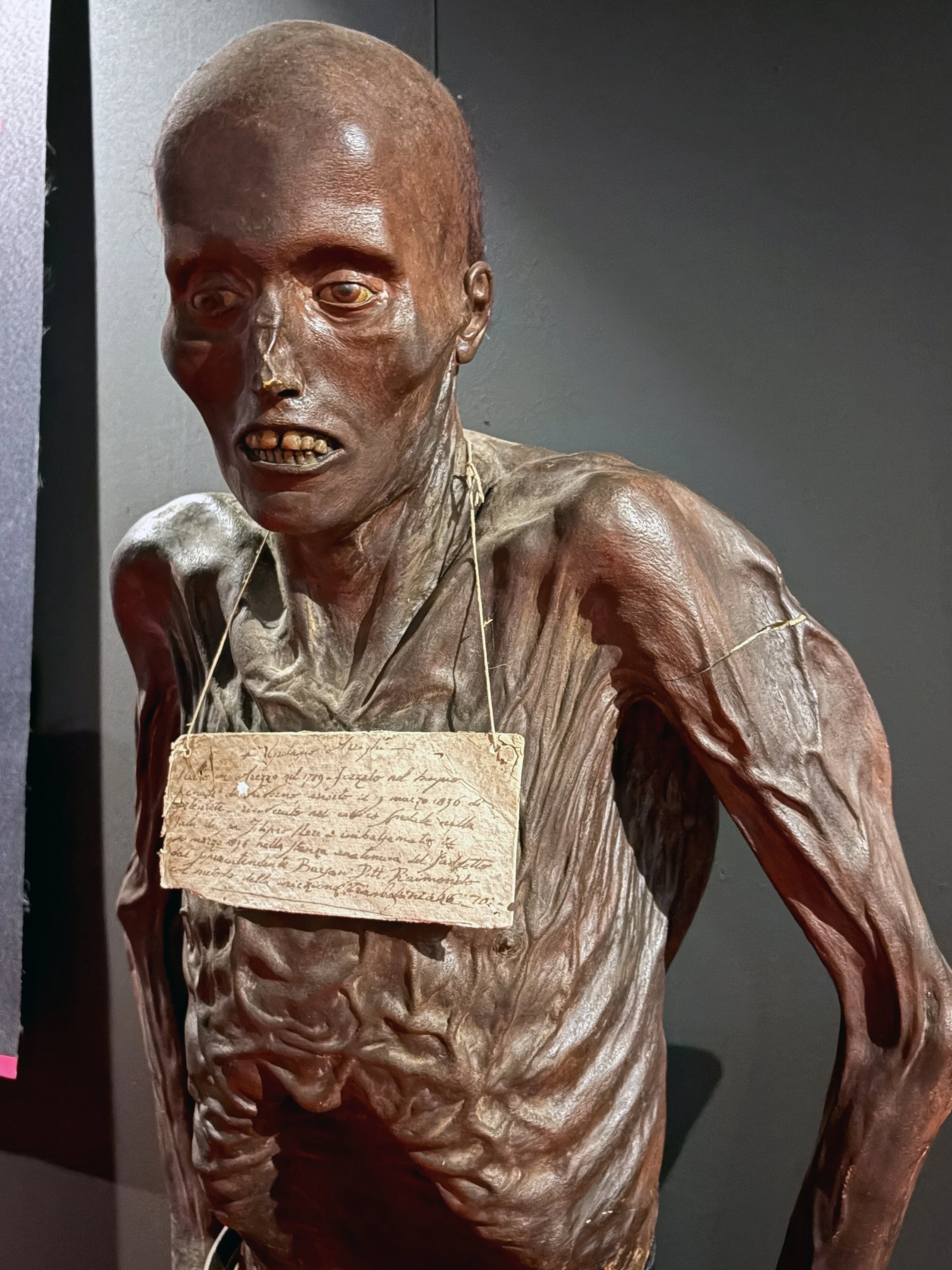 A sculpture of a human figure with exaggerated features, including a large head, prominent cheekbones, and an open mouth showing uneven teeth, with a writing note hanging around its neck.