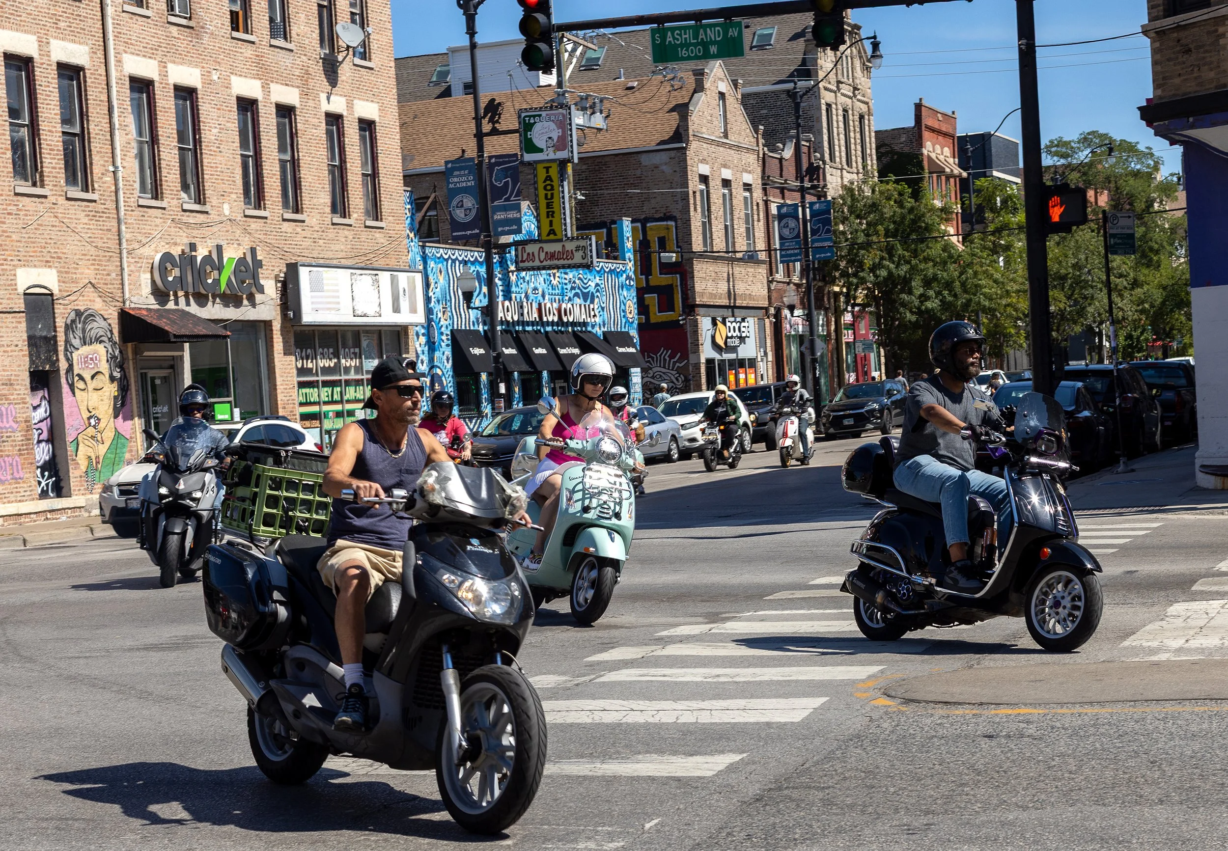Multiple motorcyclists and scooters crossing an intersection in an urban area with brick buildings, storefronts, and signage, including a Cricket store and signs indicating South Ashland Avenue.