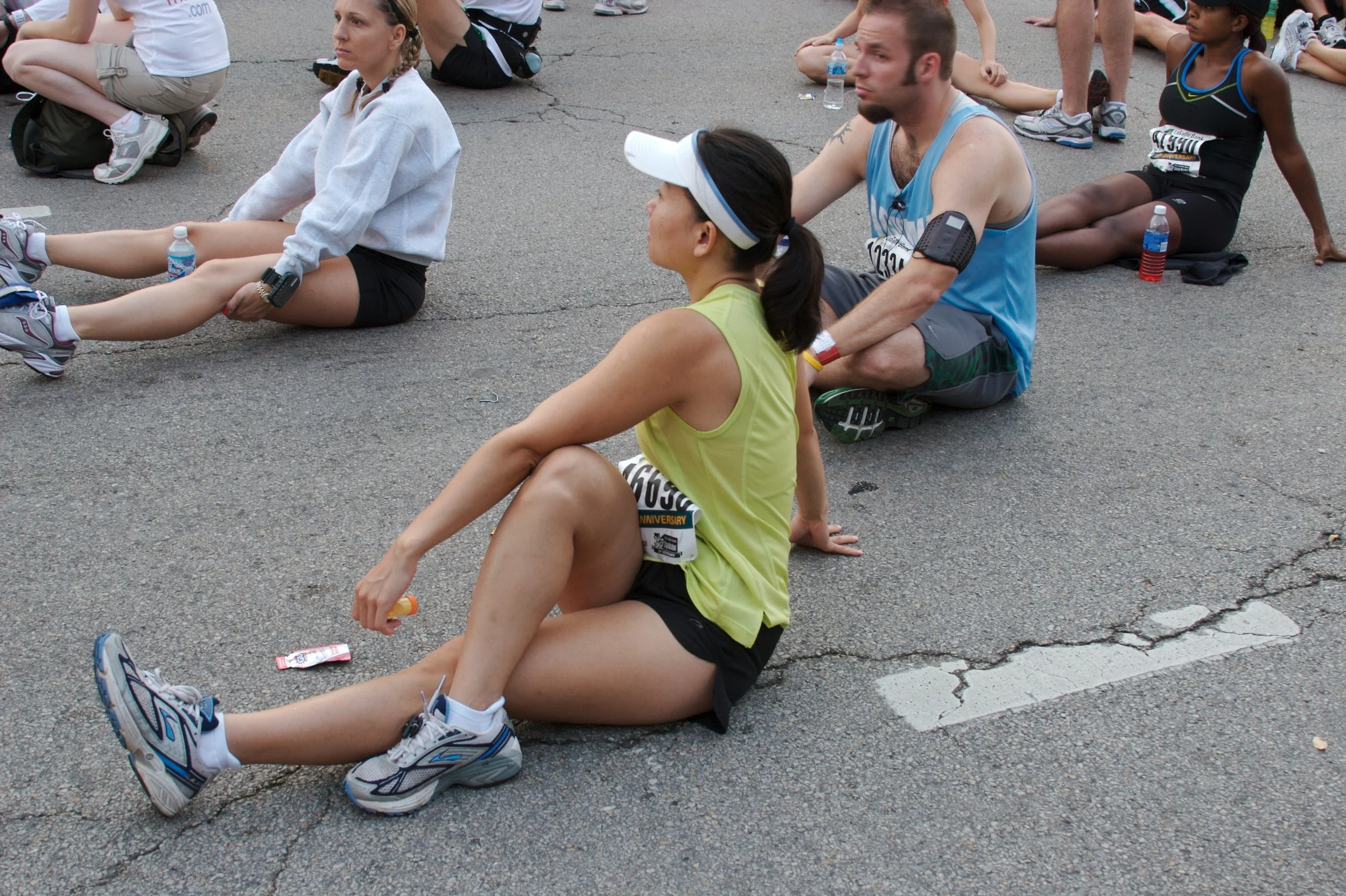 A group of people sitting on the pavement, stretching and resting during a running event. They are wearing athletic clothing, some with race bibs, and have water bottles nearby.