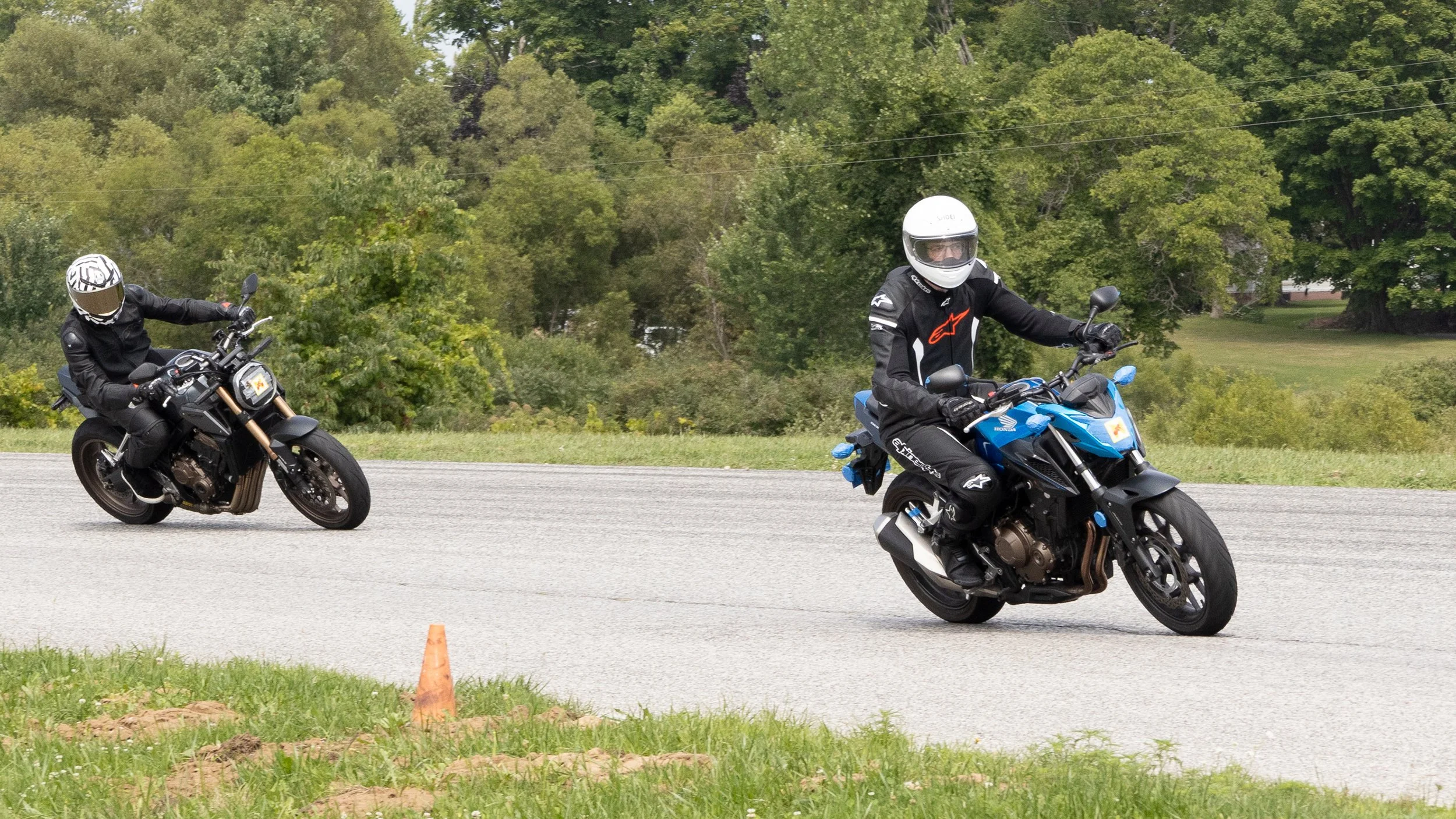 Two motorcyclists riding on a paved road, wearing helmets and protective gear, with green trees and a grassy background.