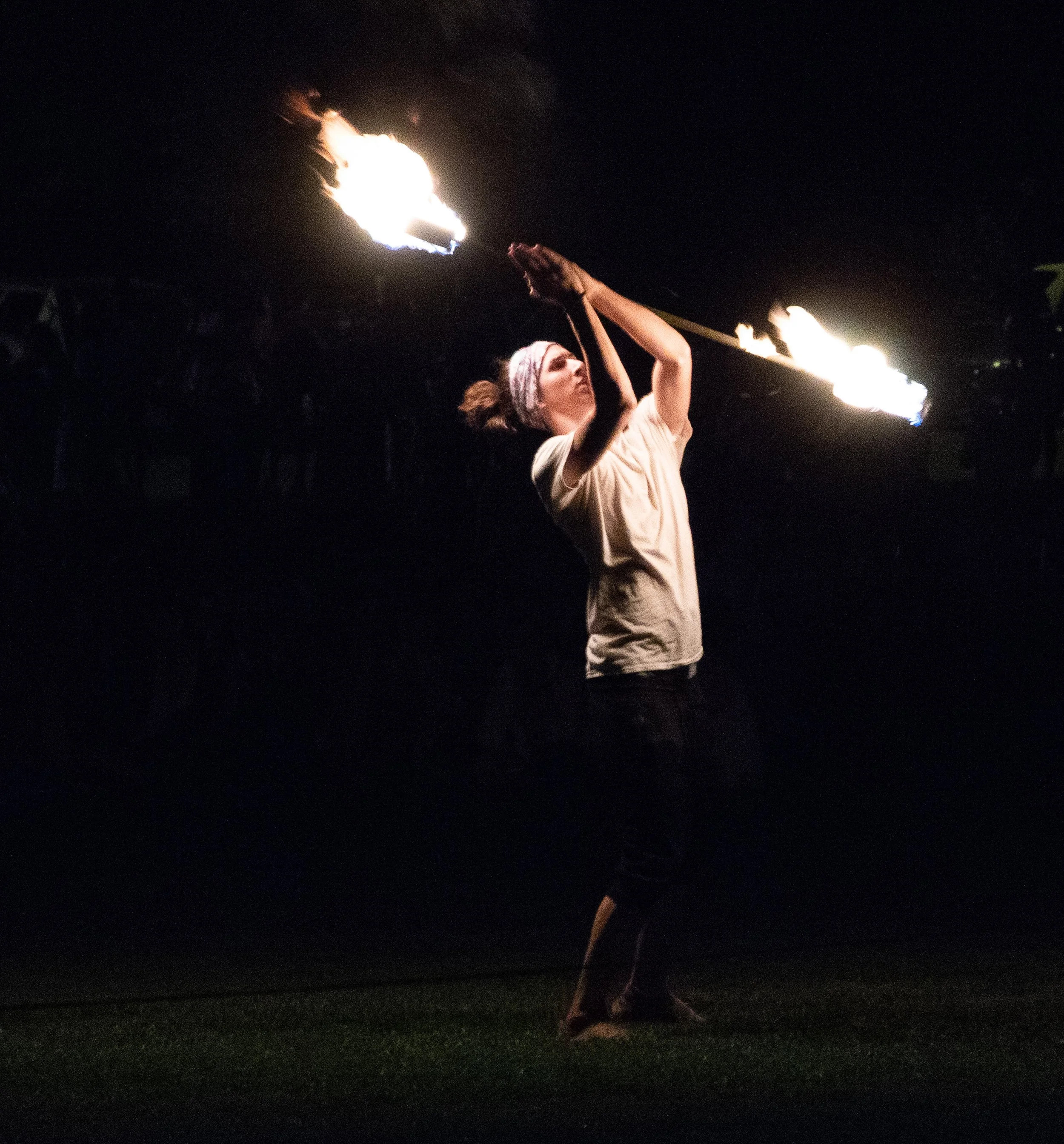 A woman in a light-colored shirt and headband holds a flaming torch at night, with a dark background.