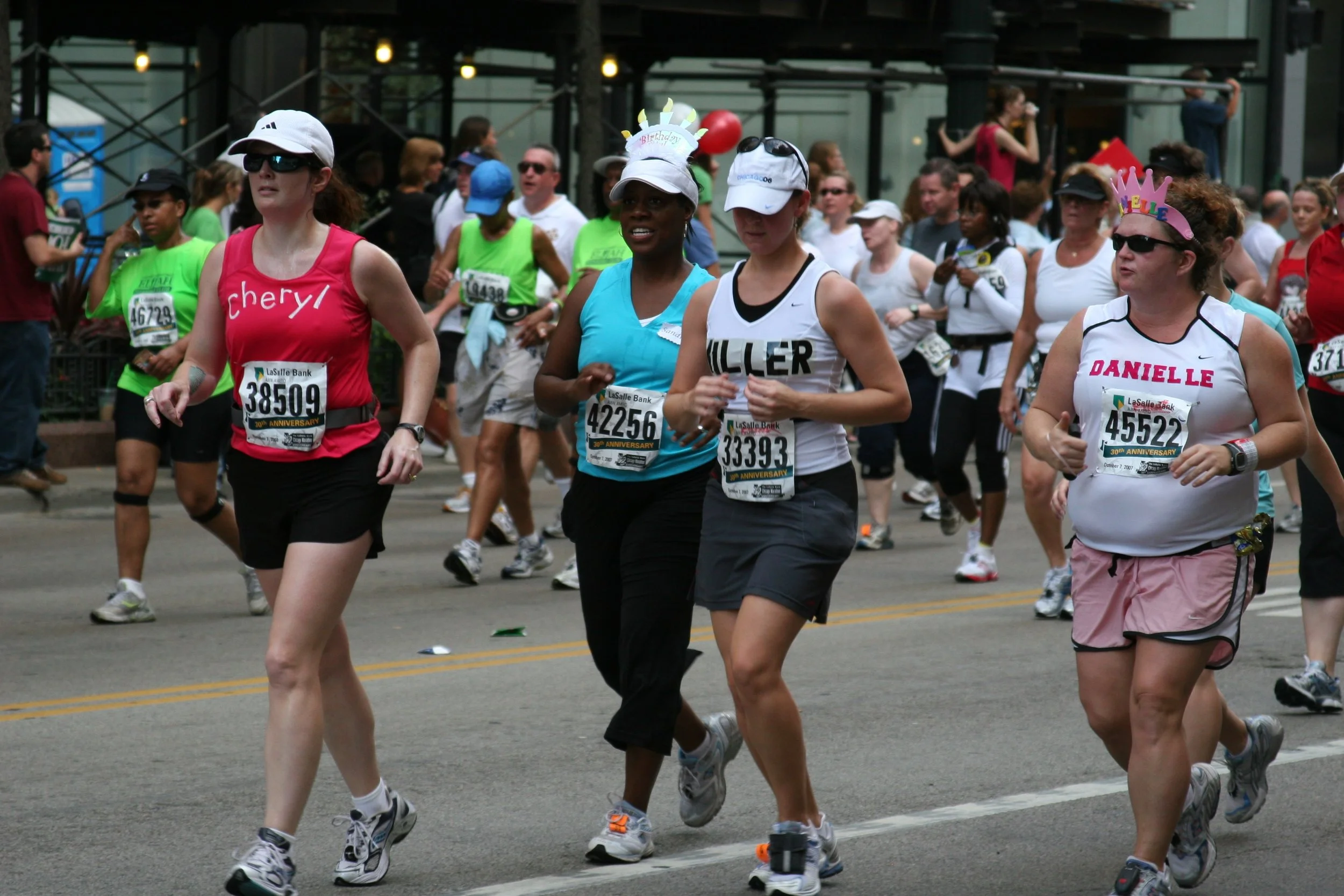 A group of runners participating in a marathon or race, some wearing bib numbers and hats, running on a city street with spectators watching.