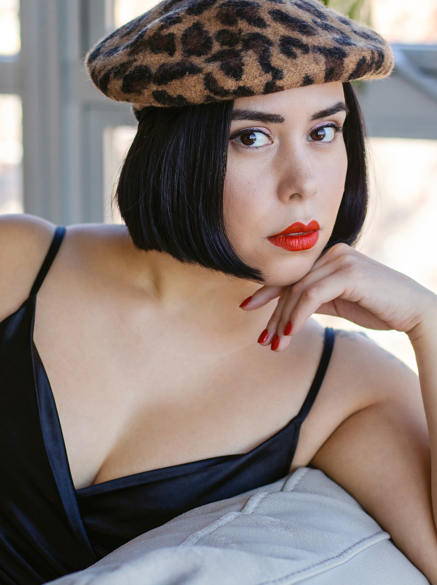 A woman with short black hair wearing a leopard print beret, red lipstick, and a black spaghetti strap top, resting her chin on her hand.