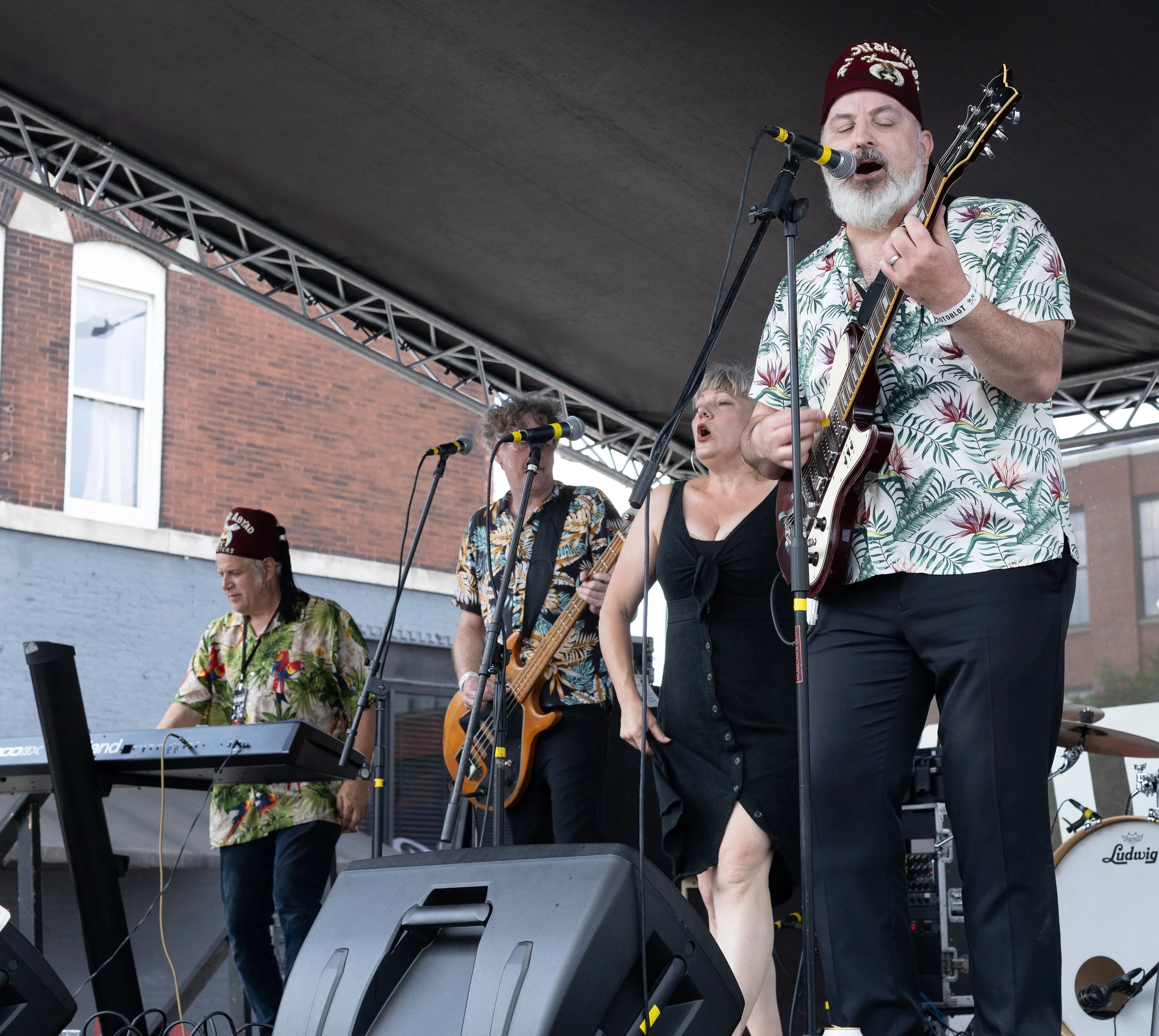 A man with a gray beard wearing a patterned hawaiian shirt and a red hat playing an electric guitar with eyes closed on an outdoor stage. Other musicians are present, one playing keyboard, in front of a brick building.