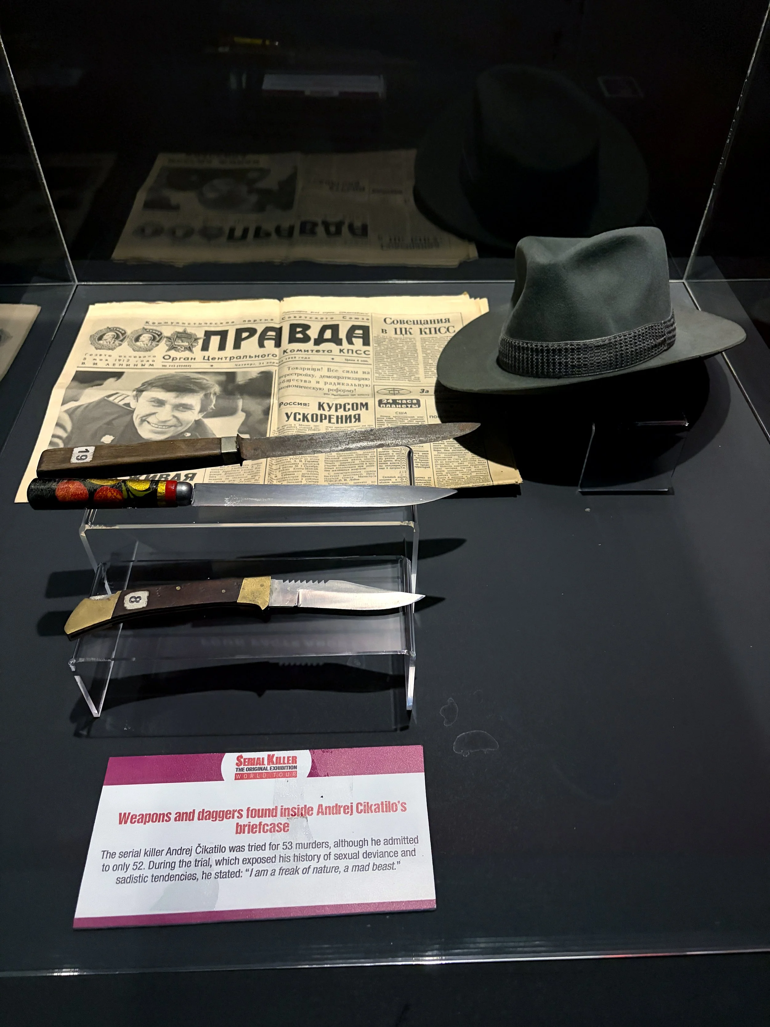 Display case containing two guns, a newspaper, a hat, and two knives with a museum informational placard explaining they are weapons and daggers found inside the briefcase of serial killer Andrei Cikatilo, including details about his crimes and psych