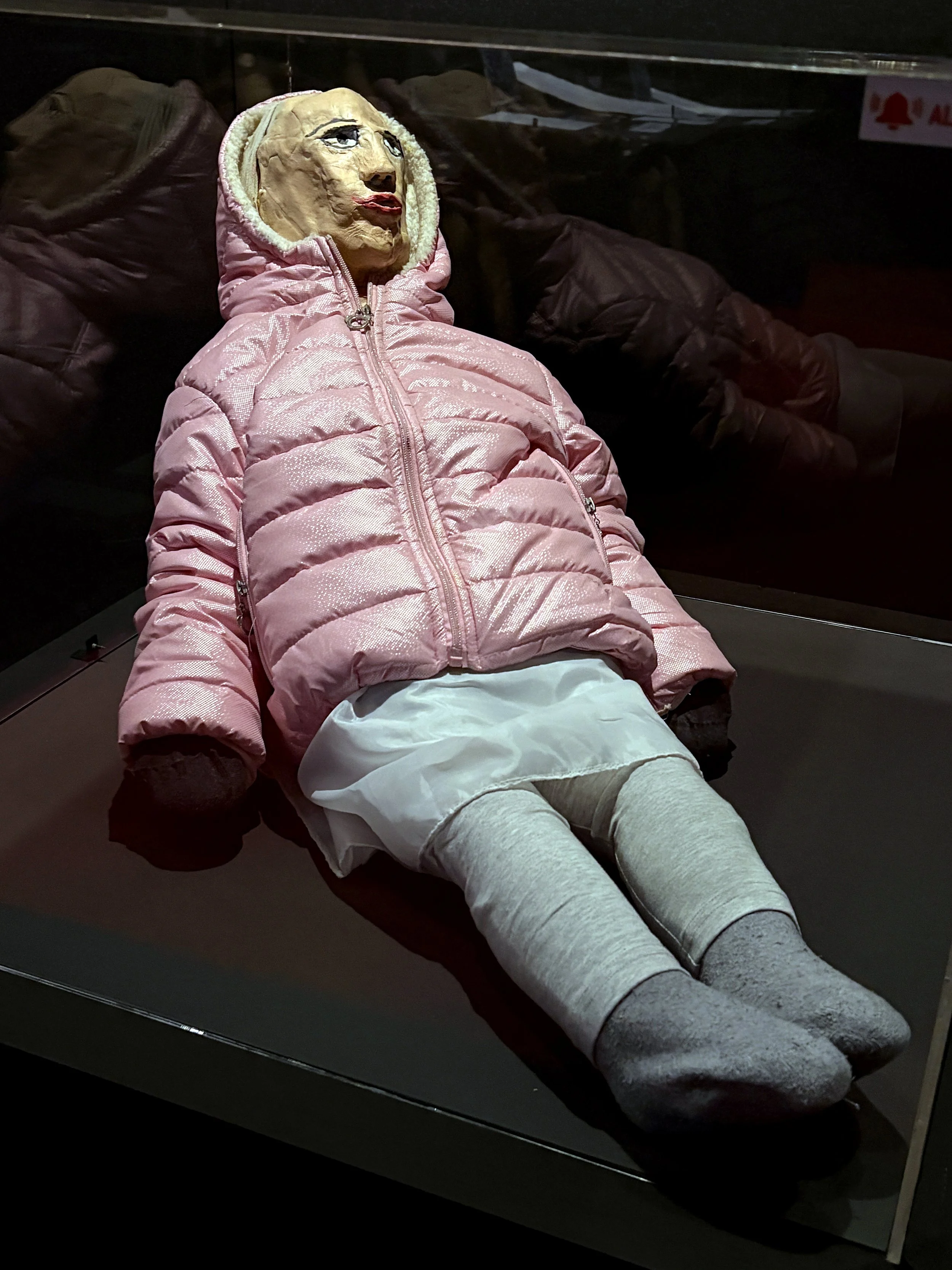 A mannequin with a human-like face wearing a pink puffy jacket and grey leggings, lying on a display table in a museum or exhibit setting.