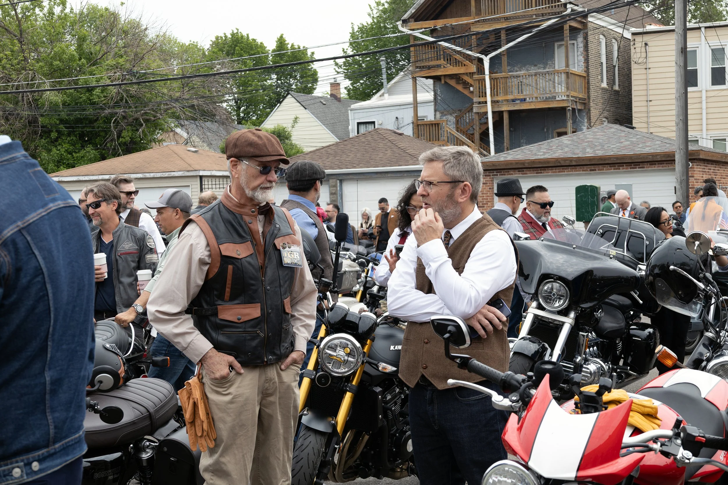 People gathered around motorcycles in a residential neighborhood with houses and trees in the background.