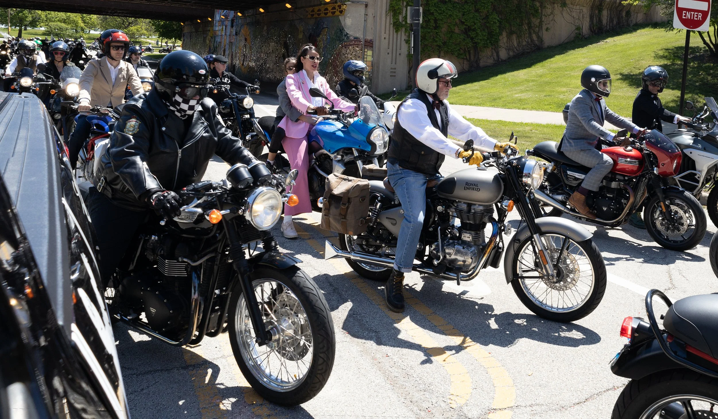 A group of people riding motorcycles and scooters through an underpass on a sunny day, with some wearing helmets and stylish clothing.