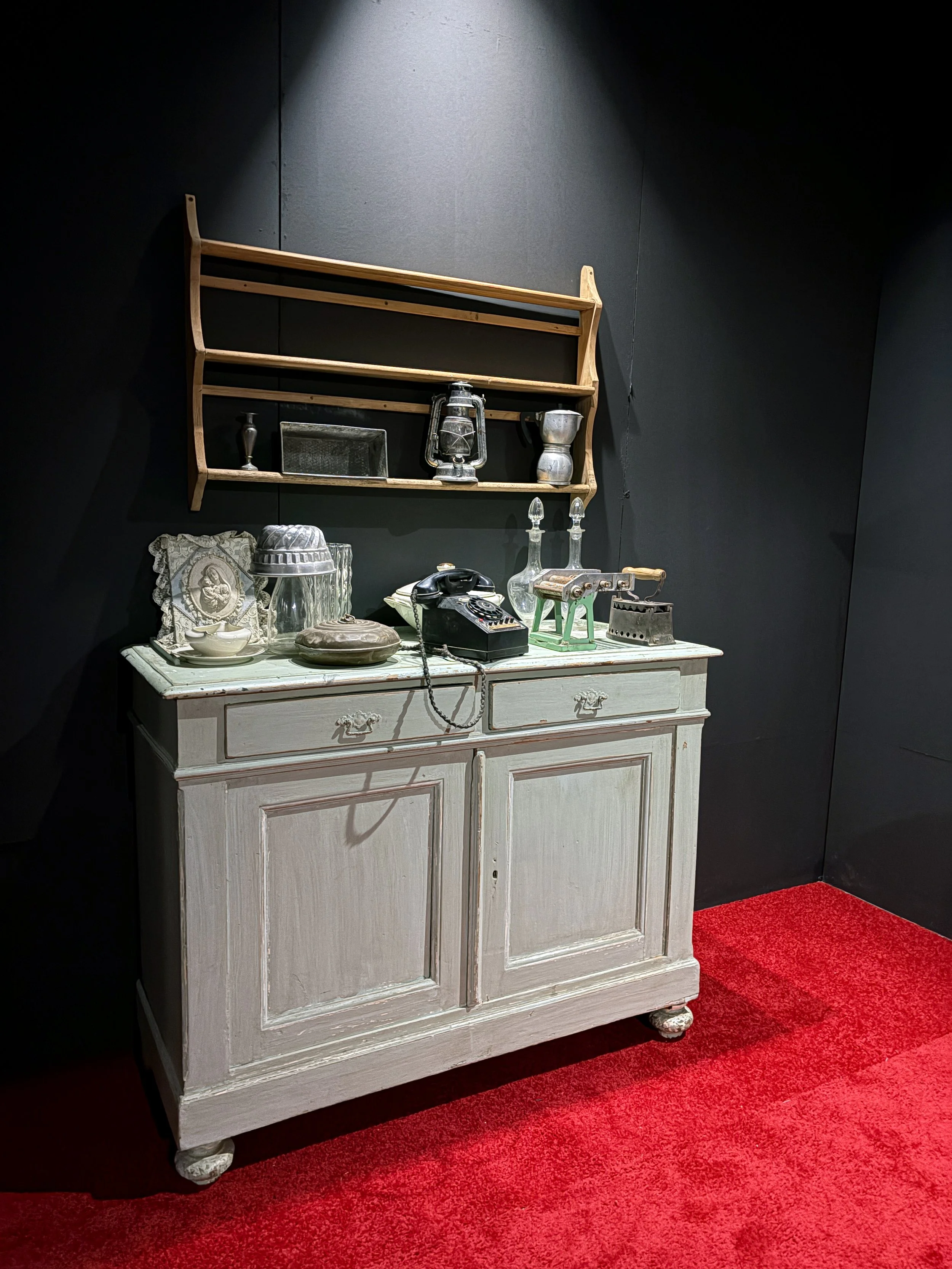 Vintage white wooden dresser with decorative carvings on the corners, placed against a black wall. On top, there are glassware, an old-fashioned black rotary telephone, and various decorative objects. A wooden wall shelf above stores a lantern, silve