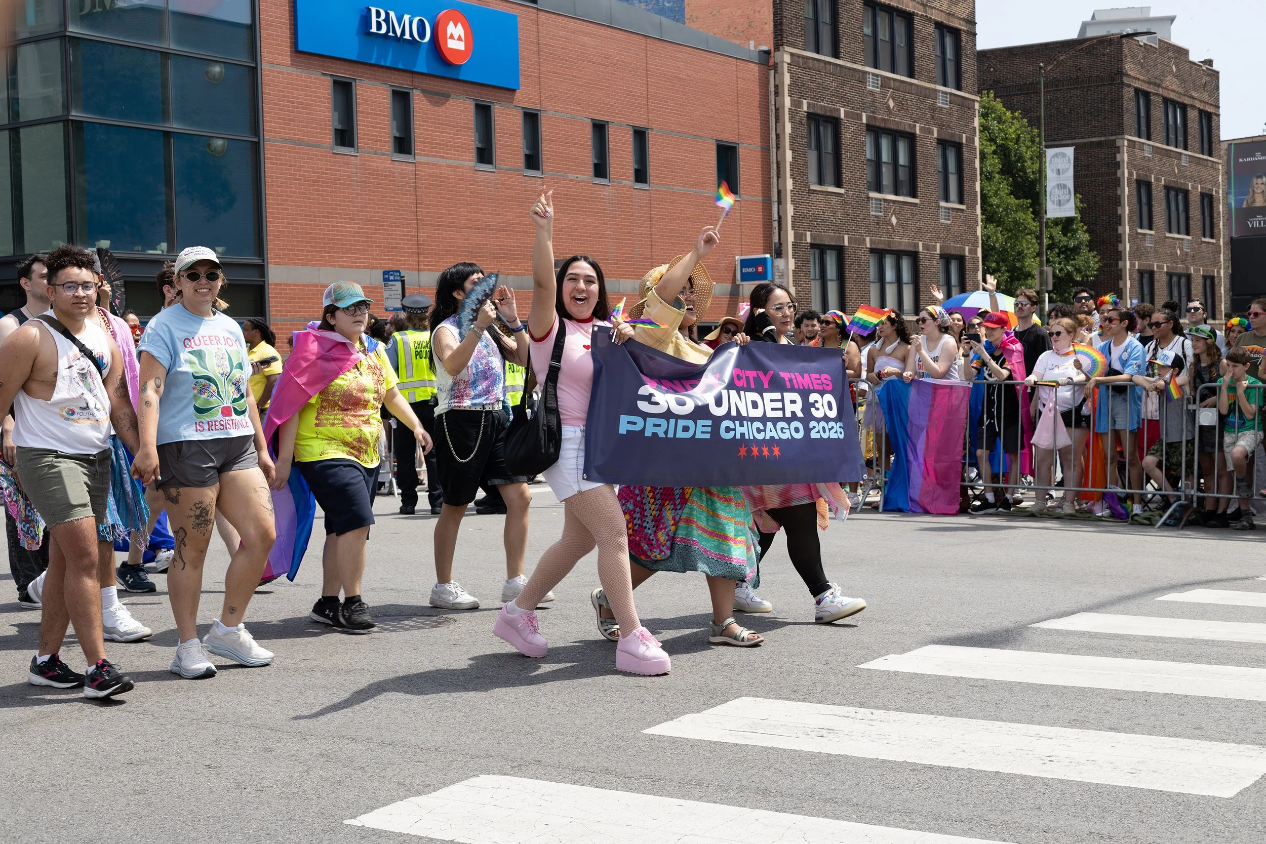 A diverse group of people marching in a Pride parade, holding a banner for a youth pride group, with some dressed in rainbow-themed clothing and accessories, on a city street near a BMO bank.